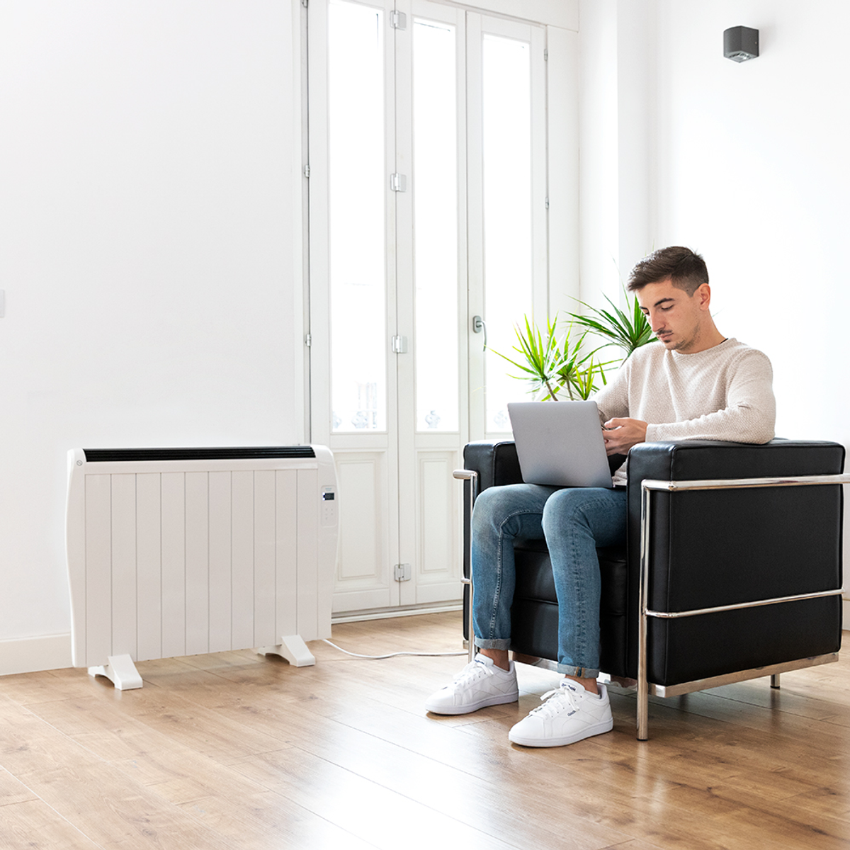 Mężczyzna w salonie używa laptopa, biały grzejnik, czarny fotel, drewniana podłoga.