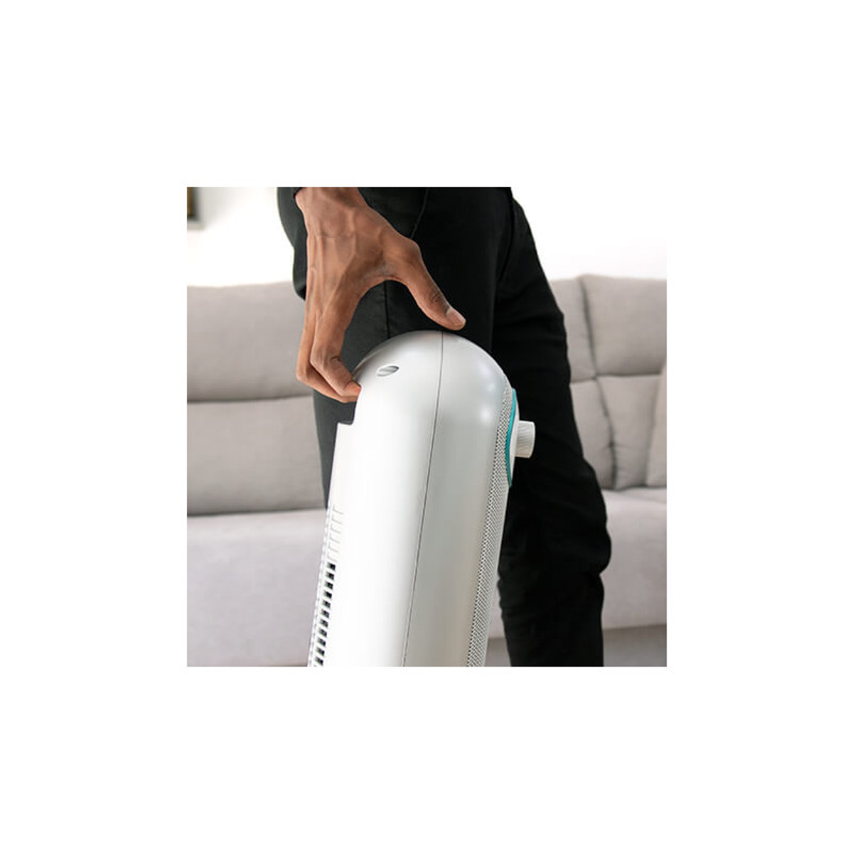 Ręka osoby dotyka górnej części białego, cylindrycznego wentylatora. Tłem jest jasnoszara sofa.