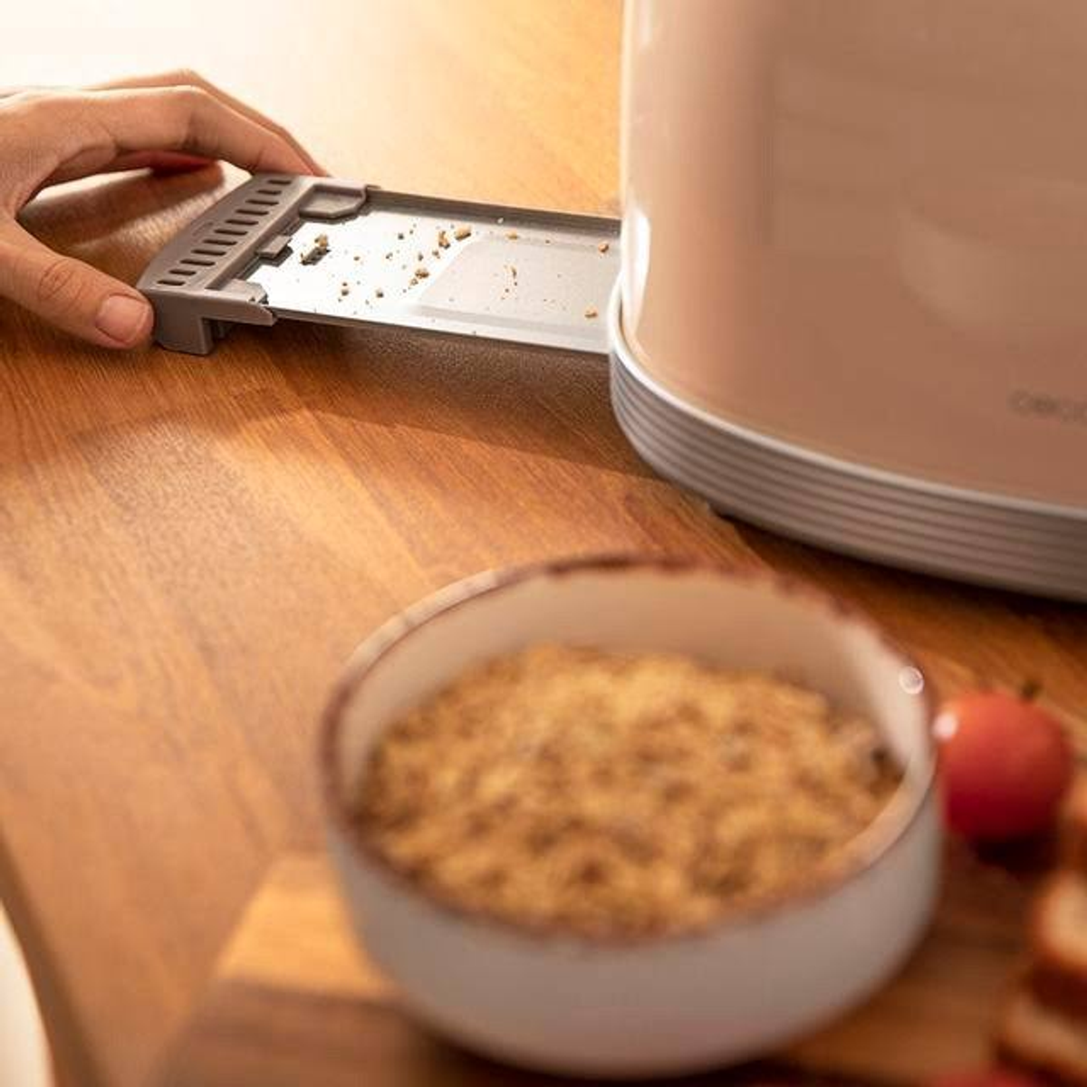 Eine Hand zieht ein Tablett eines beigen Toasters heraus. Brotkrumen sind sichtbar. Eine Schüssel mit Krümeln und Apfel ist im Hintergrund.