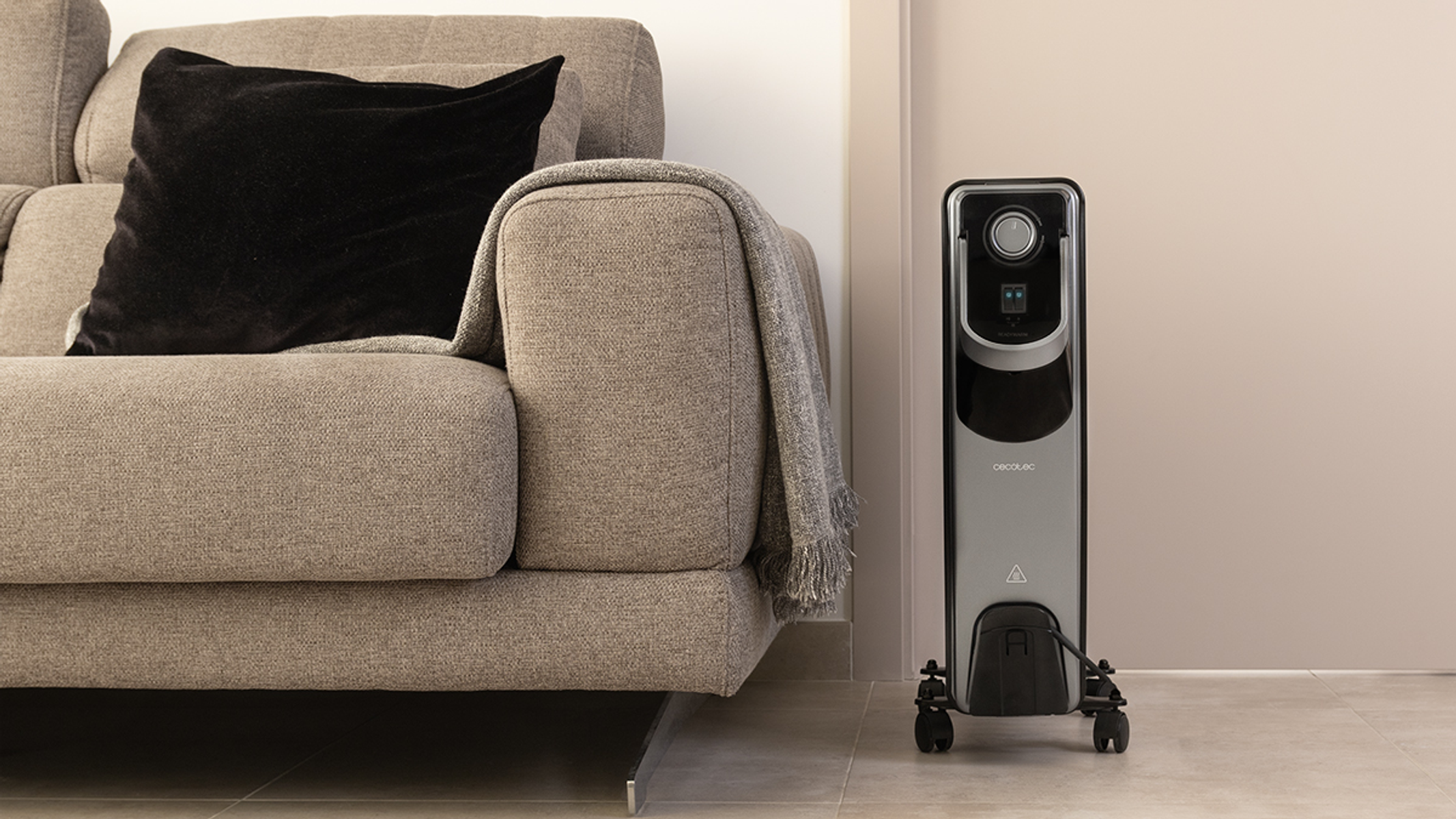 Czarny i srebrny grzejnik elektryczny obok szarej kanapy. Czarna poduszka i koc na kanapie.