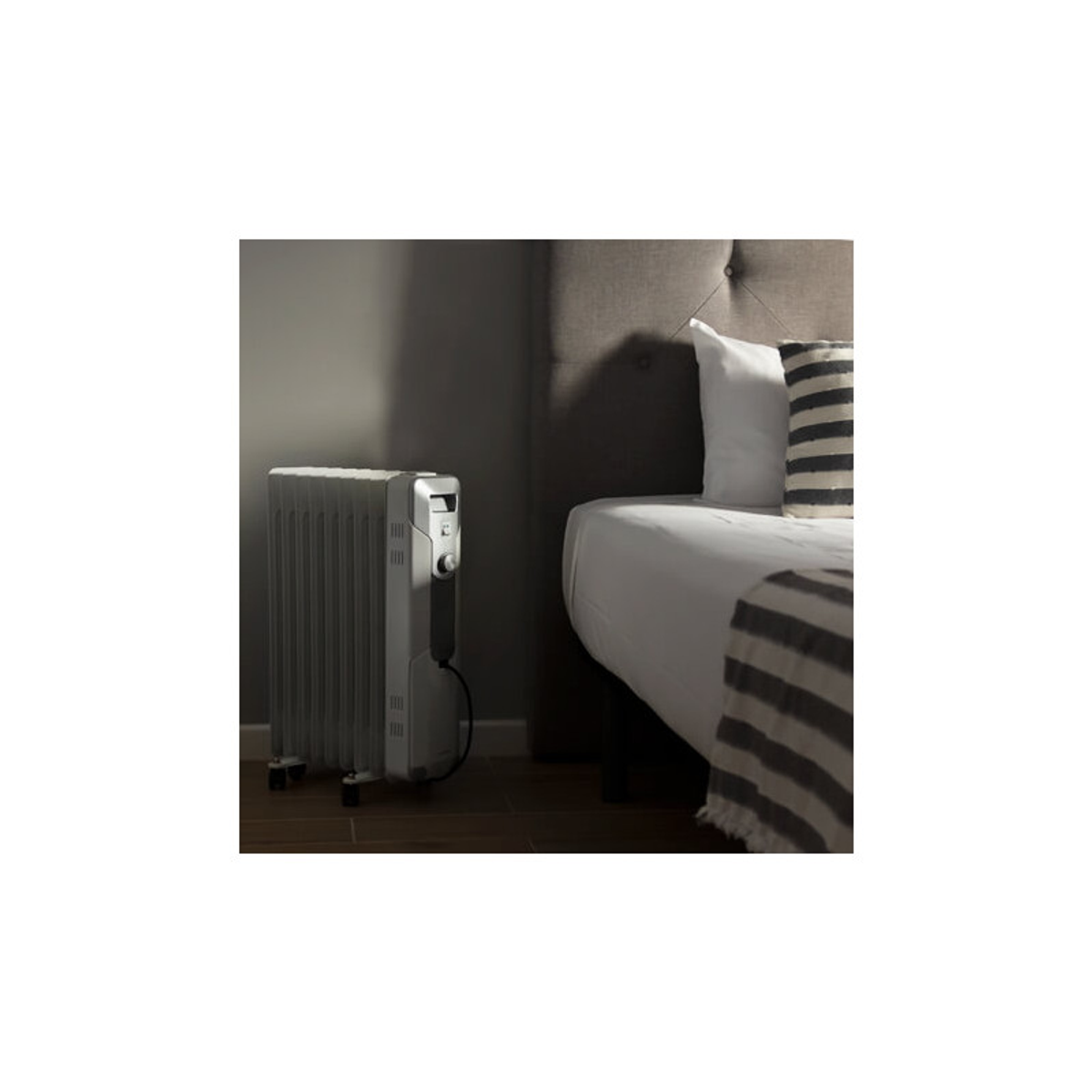 Biały grzejnik olejowy na kółkach przed łóżkiem.