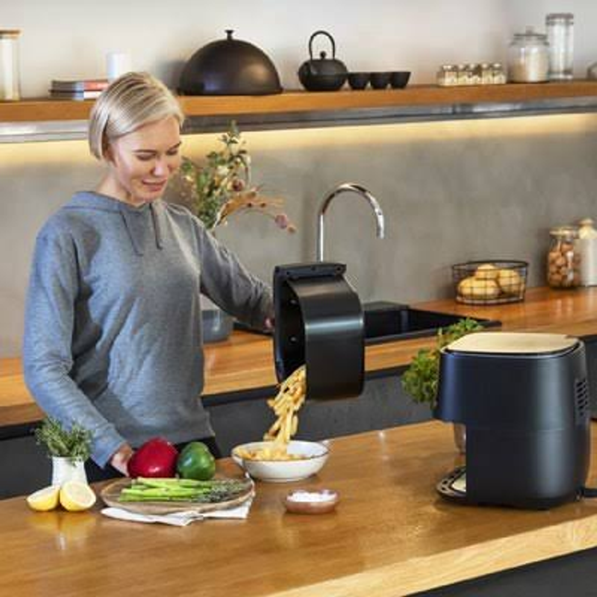 Una donna versa le patatine da una friggitrice in una ciotola, ambiente cucina con verdure.