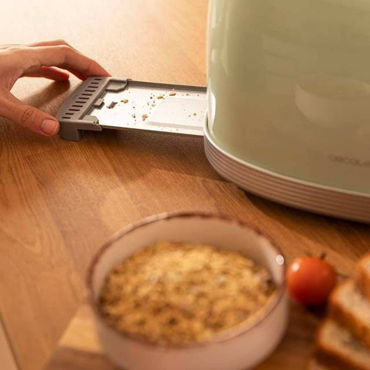 Ręka trzyma tackę na okruchy zielonego tostera, z okruchami w misce i kromkami chleba.