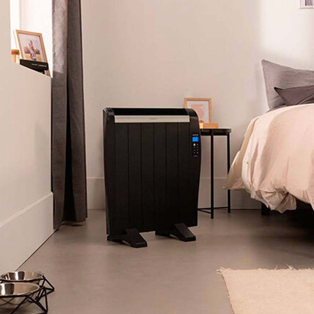 Czarny grzejnik elektryczny stojący na szarej podłodze, w pokoju z łóżkiem i oknem.