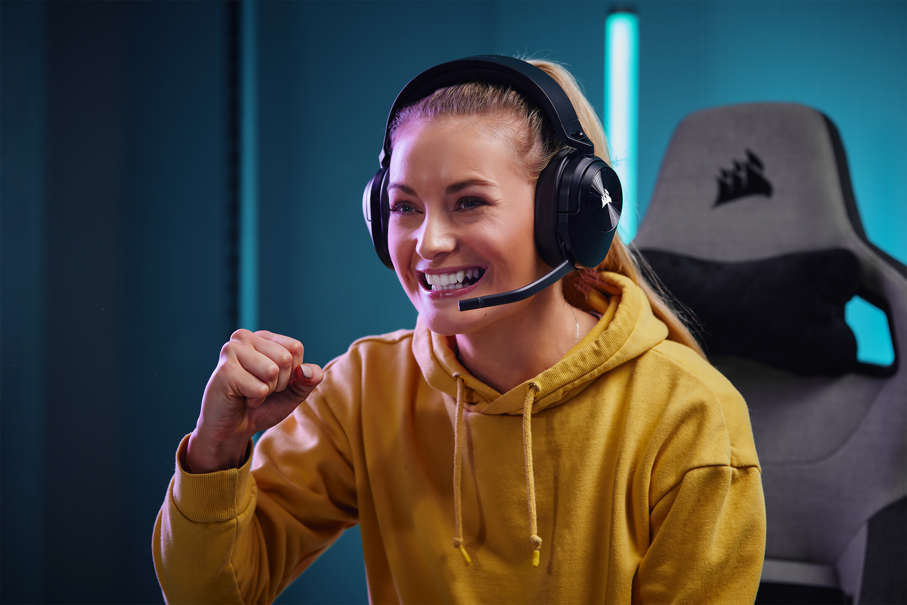 Donna con cuffie e felpa gialla, sorridente e che festeggia davanti a una sedia da gaming.