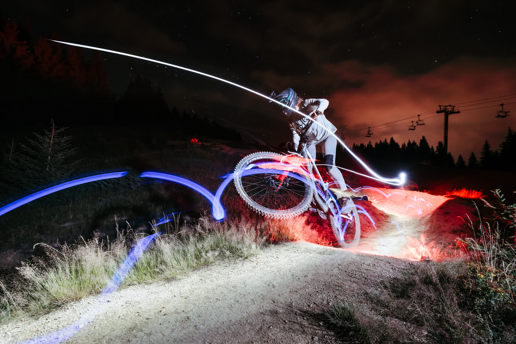 A mountain biker jumps over a dirt path at night with light trails.