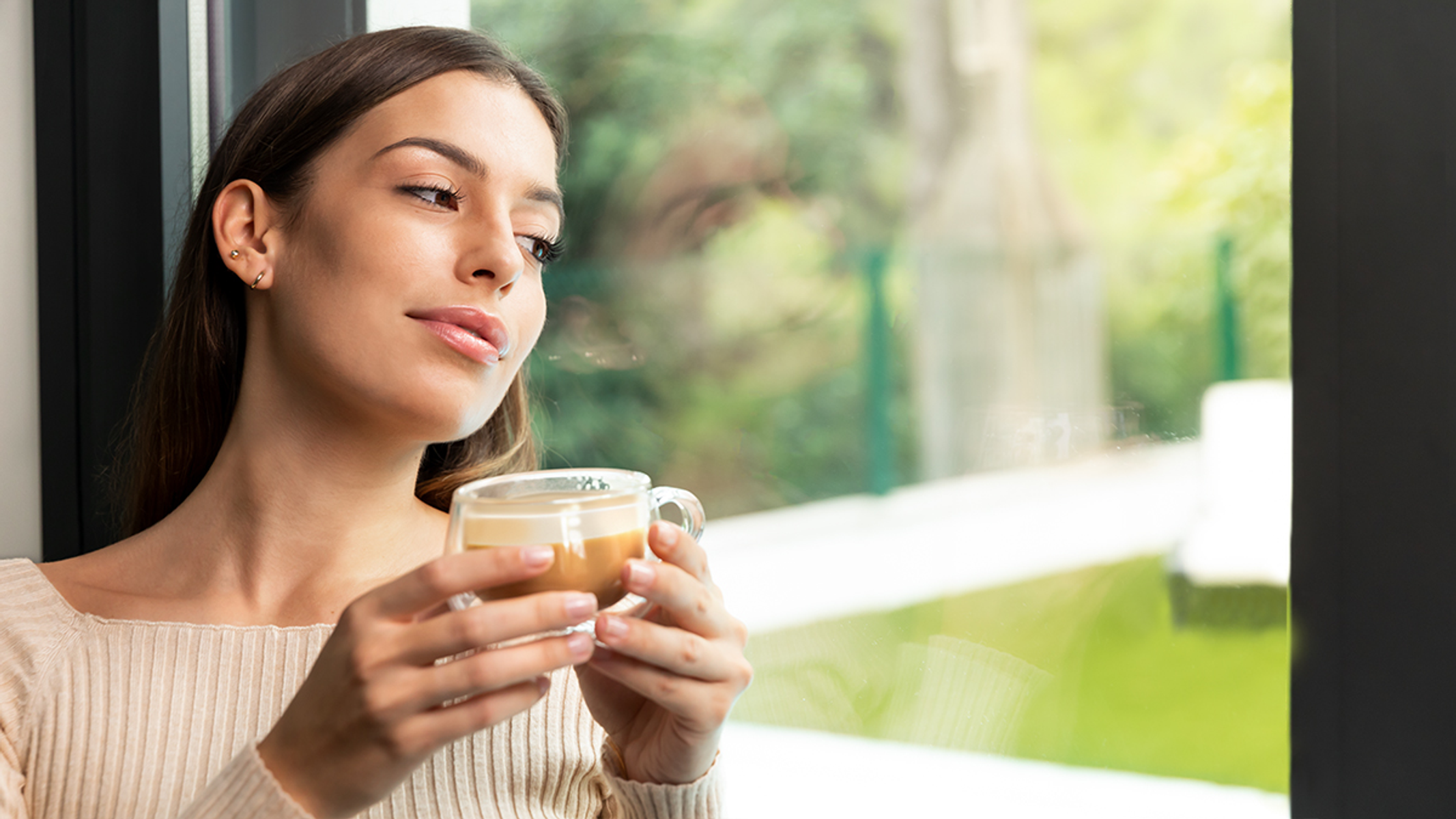Mujer con taza de café mirando por la ventana, fondo borroso.