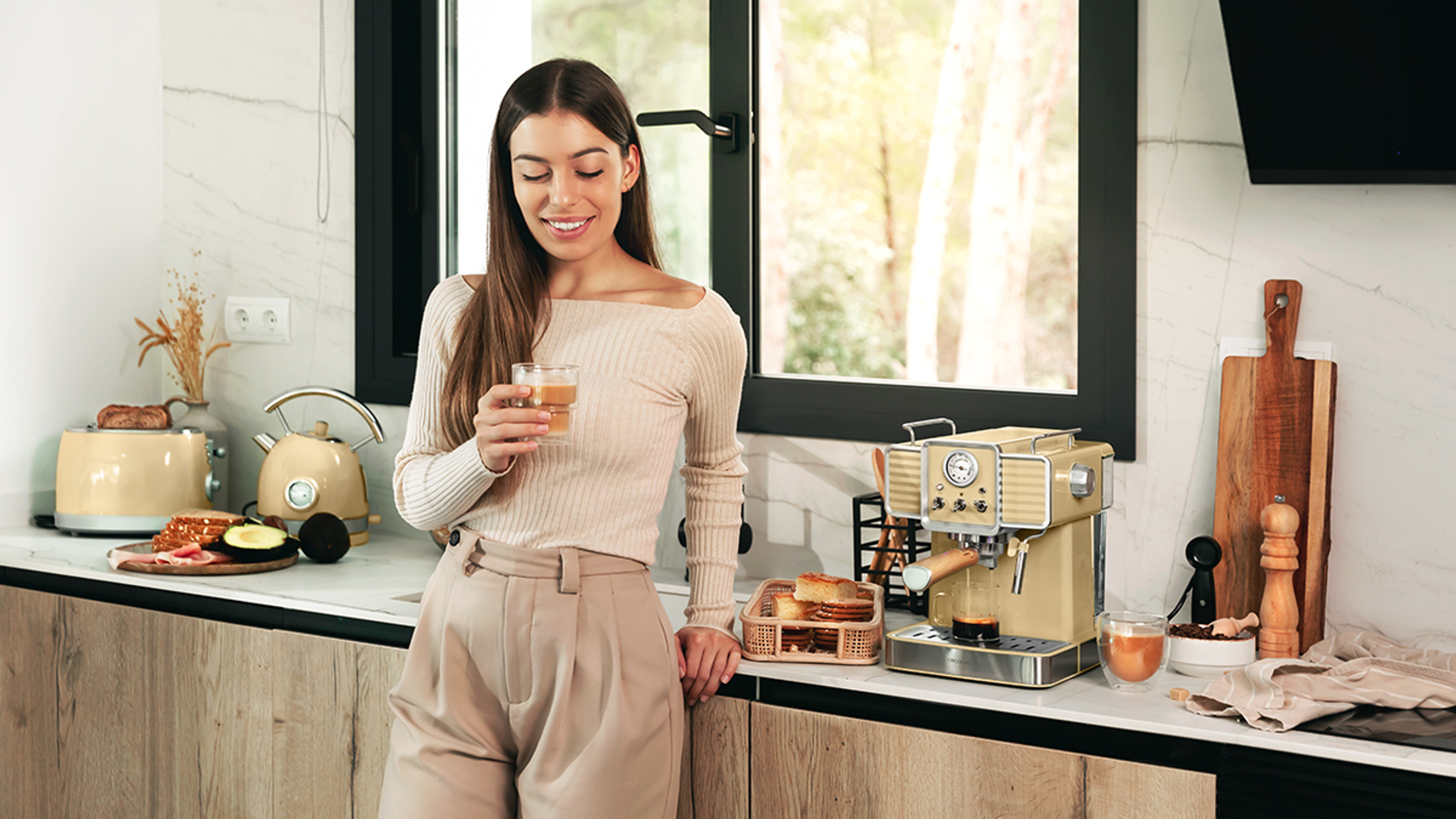 Mujer en la cocina sosteniendo un vaso de líquido, con cafetera y comida.