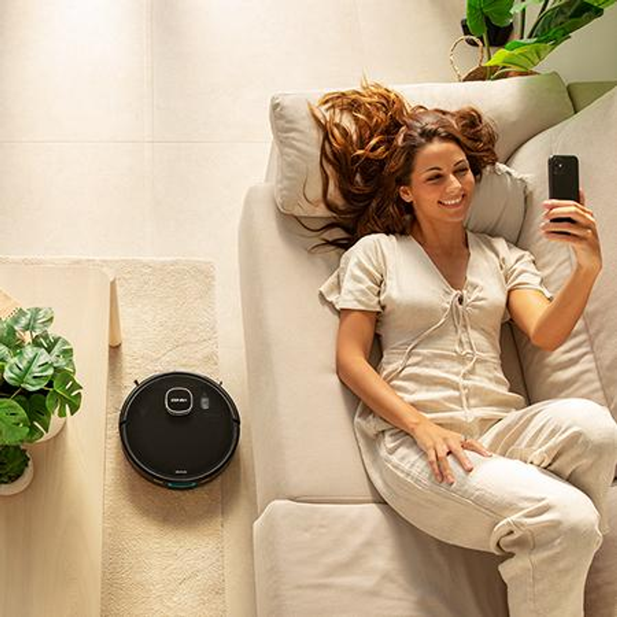 Una donna è sdraiata su un divano che si fa un selfie con un aspirapolvere robot nelle vicinanze.