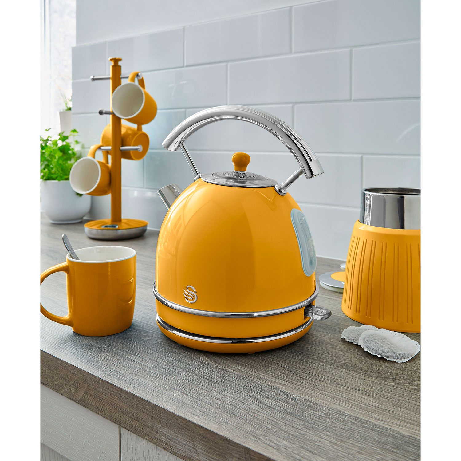 Hervidor amarillo sobre una encimera, junto a una taza, un portavasos y un infusor de té.