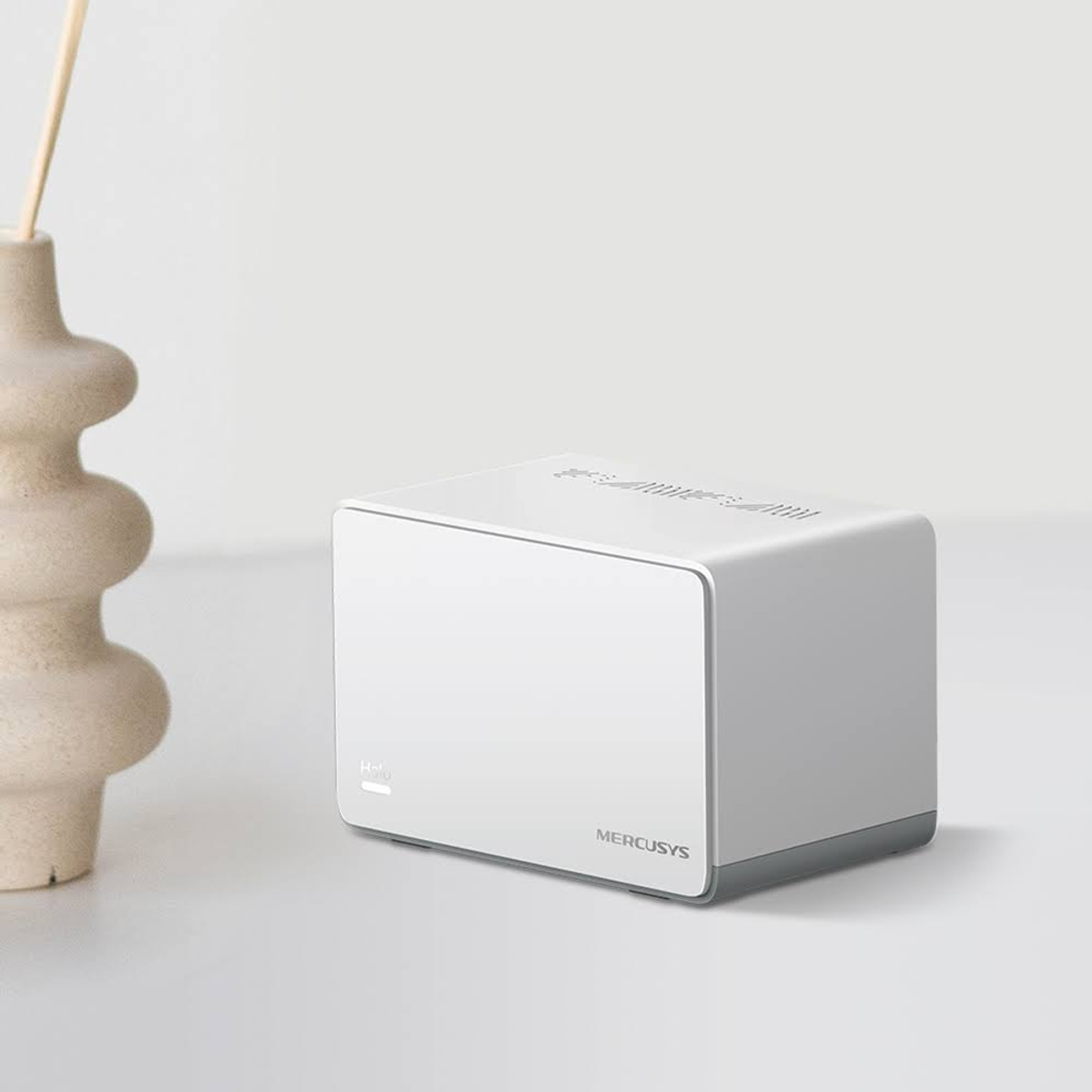 Biały prostokątny router Mercusys. Wazon z rośliną po lewej stronie routera.