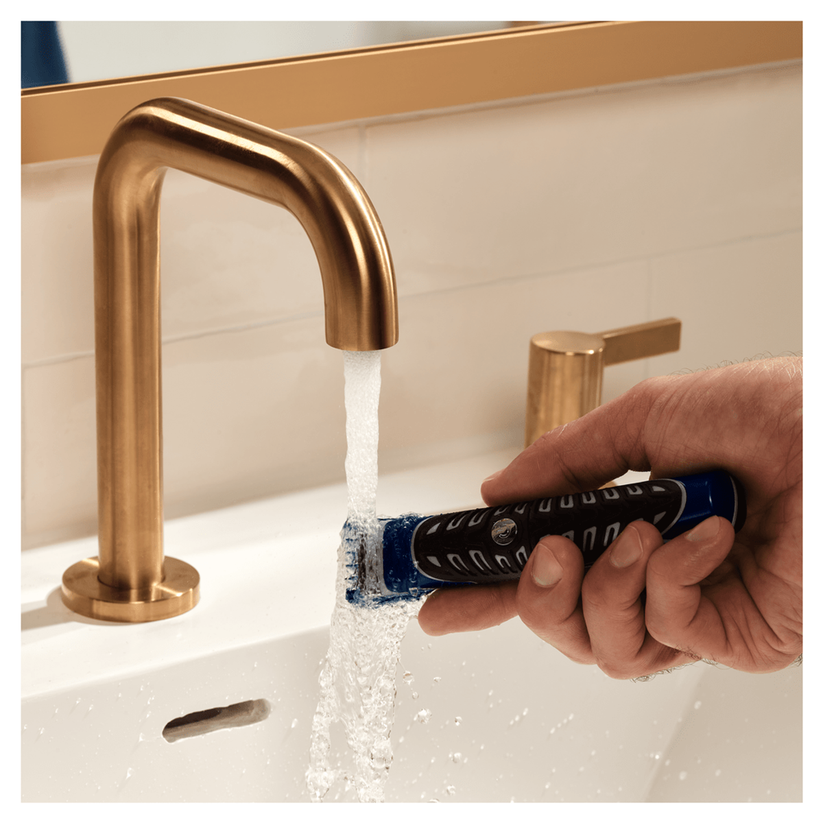 Una mano usando una afeitadora eléctrica azul y negra bajo un chorro de agua, con un grifo dorado.
