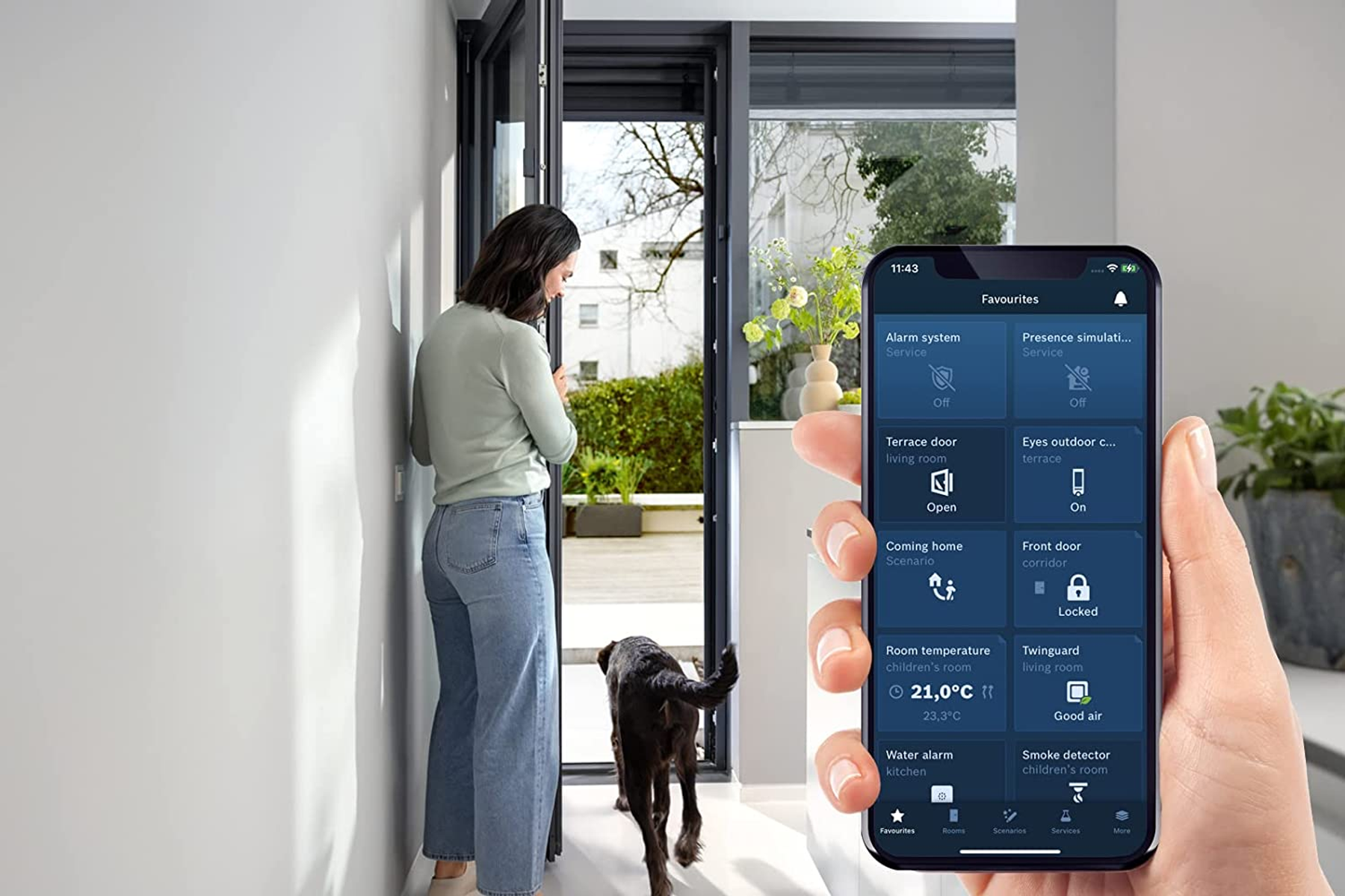 Frau schaut auf Telefon mit Smart-Home-App. Schwarzer Hund im Hintergrund. Haustür im Blick.