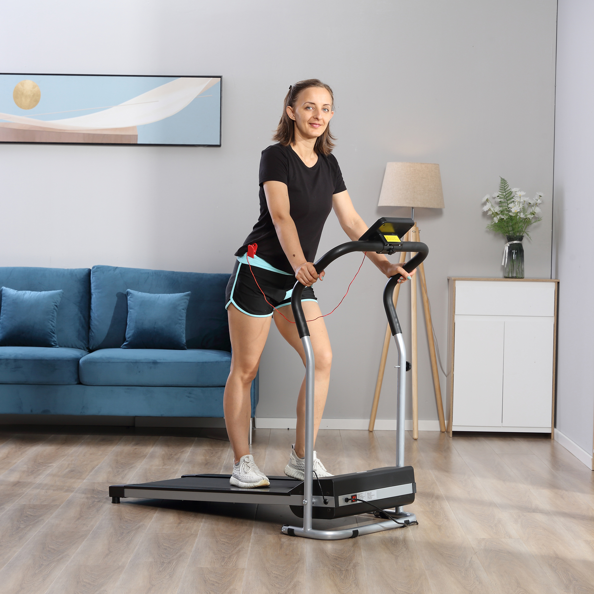 Donna su un tapis roulant in un ambiente domestico. Indossa top e pantaloncini neri. Divano blu sullo sfondo.