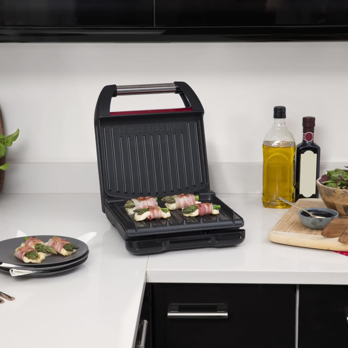 Parrilla abierta con comida cocinándose. También incluye un plato con comida y botellas en el fondo. Mostrador blanco.