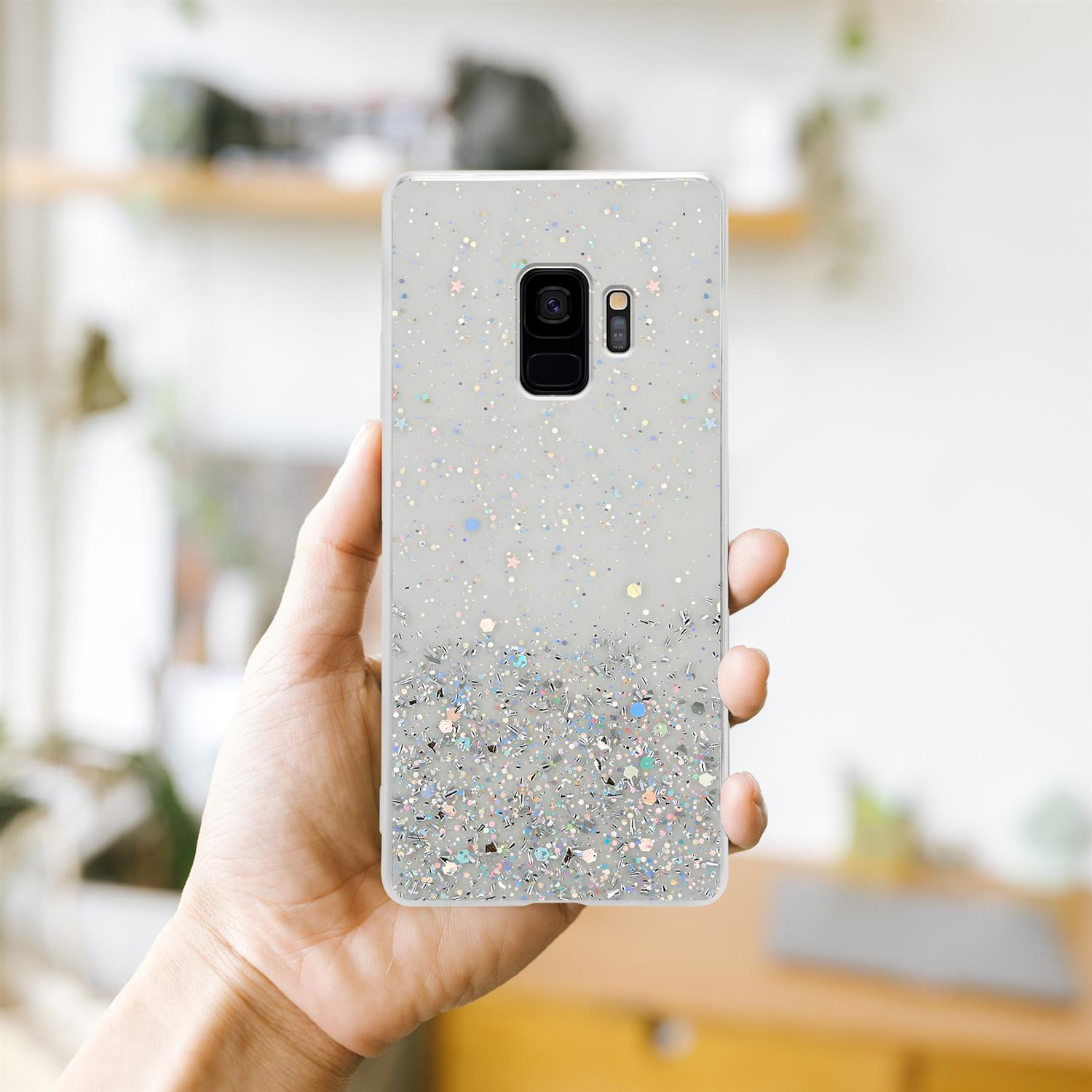 Hand hält Telefon mit glitzerndem Gehäuse. Verschwommener Hintergrund mit Regal.