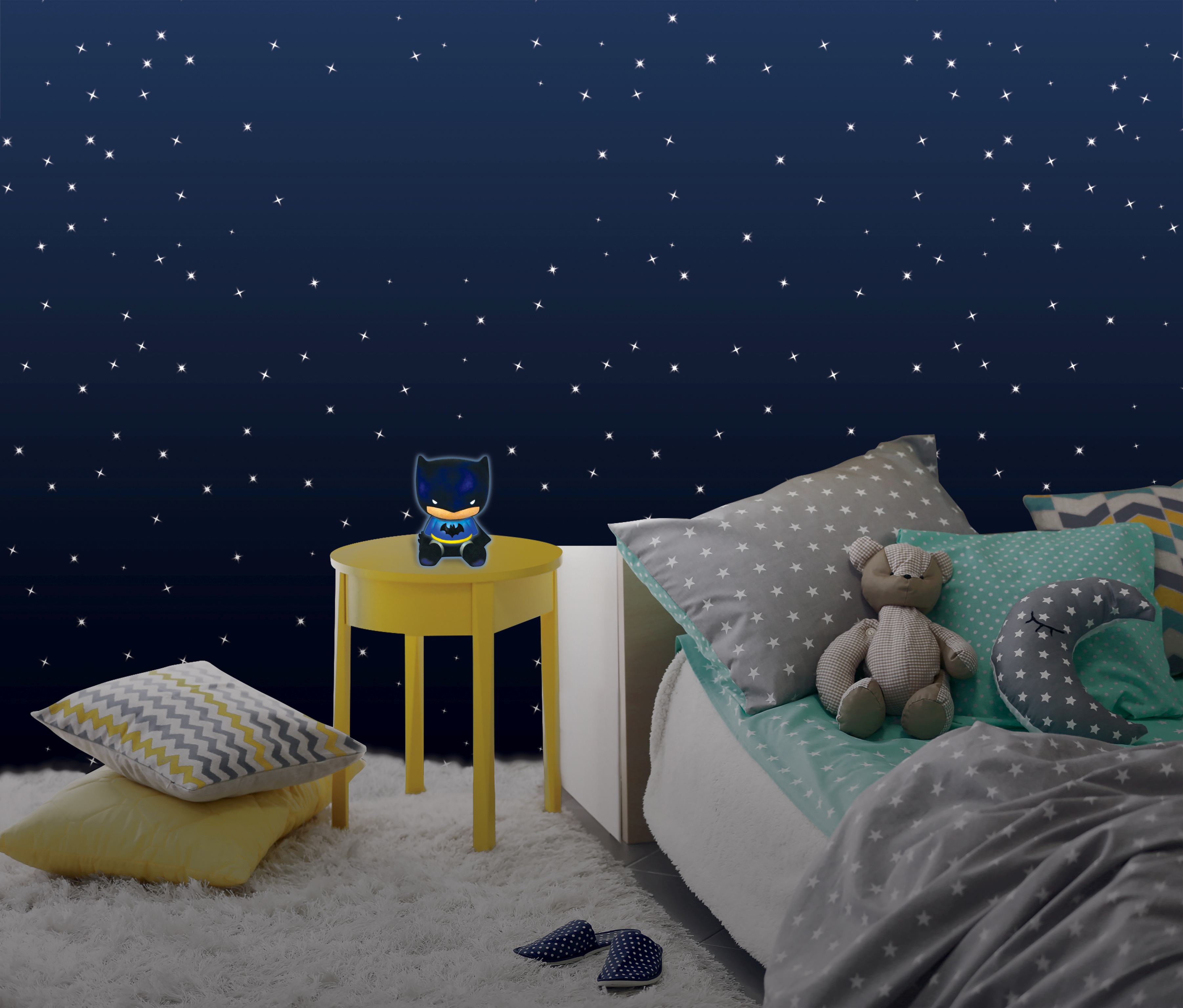 Una lámpara de Batman sobre una mesa amarilla en una habitación infantil con cama y pared de noche estrellada.