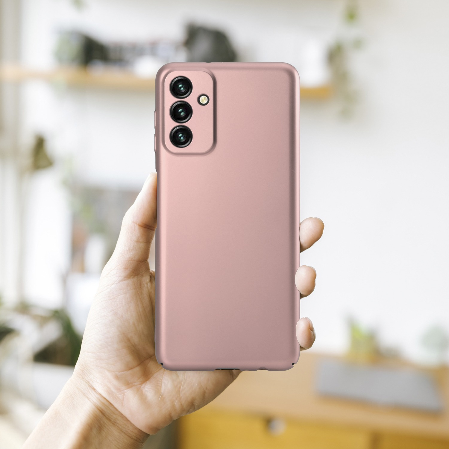 Hand hält eine rosa Handyhülle mit drei Rückkameras. Verschwommener Hintergrund eines Raumes.