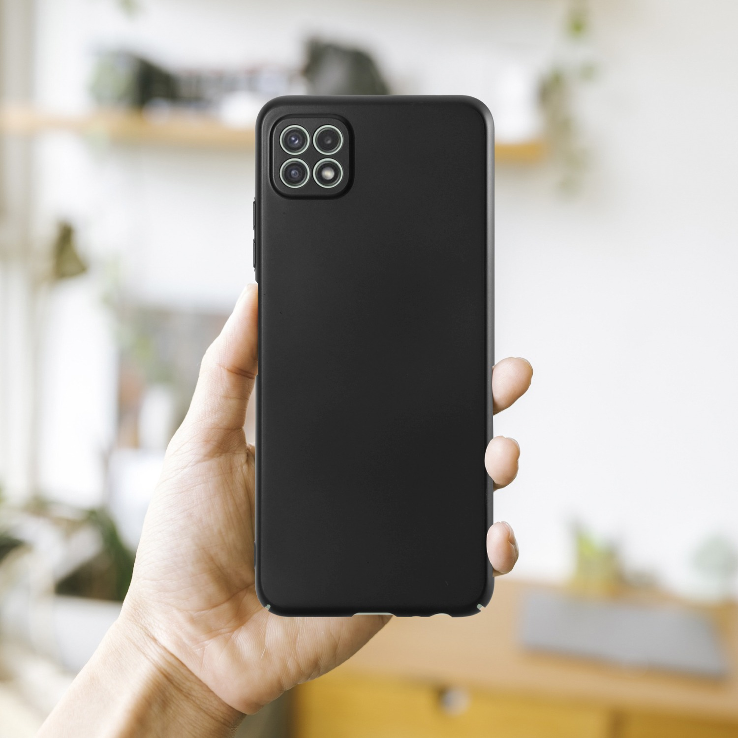 Eine Hand, die eine schwarze Handyhülle mit vier Kameras hält. Verschwommener Hintergrund mit einem Regal.