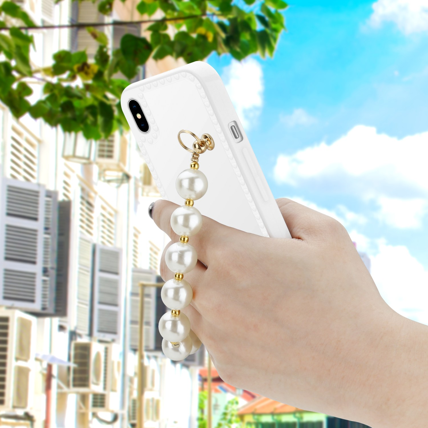 Eine Hand, die ein weißes Telefon mit einem Perlenriemen vor einem Stadtbild mit Bäumen und hellem Himmel hält.