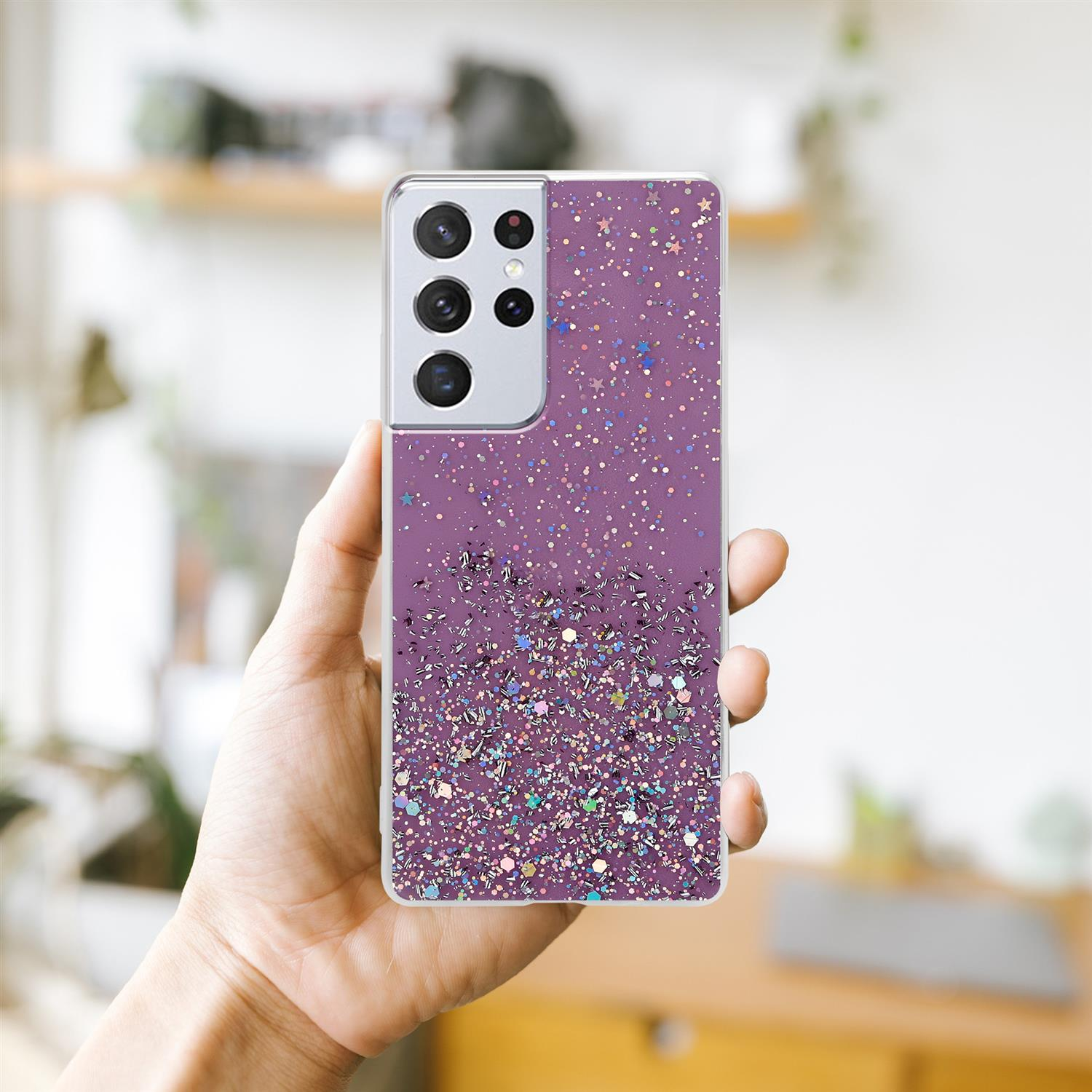Una mano tiene un telefono con una custodia viola scintillante. Lo sfondo include una mensola con oggetti.