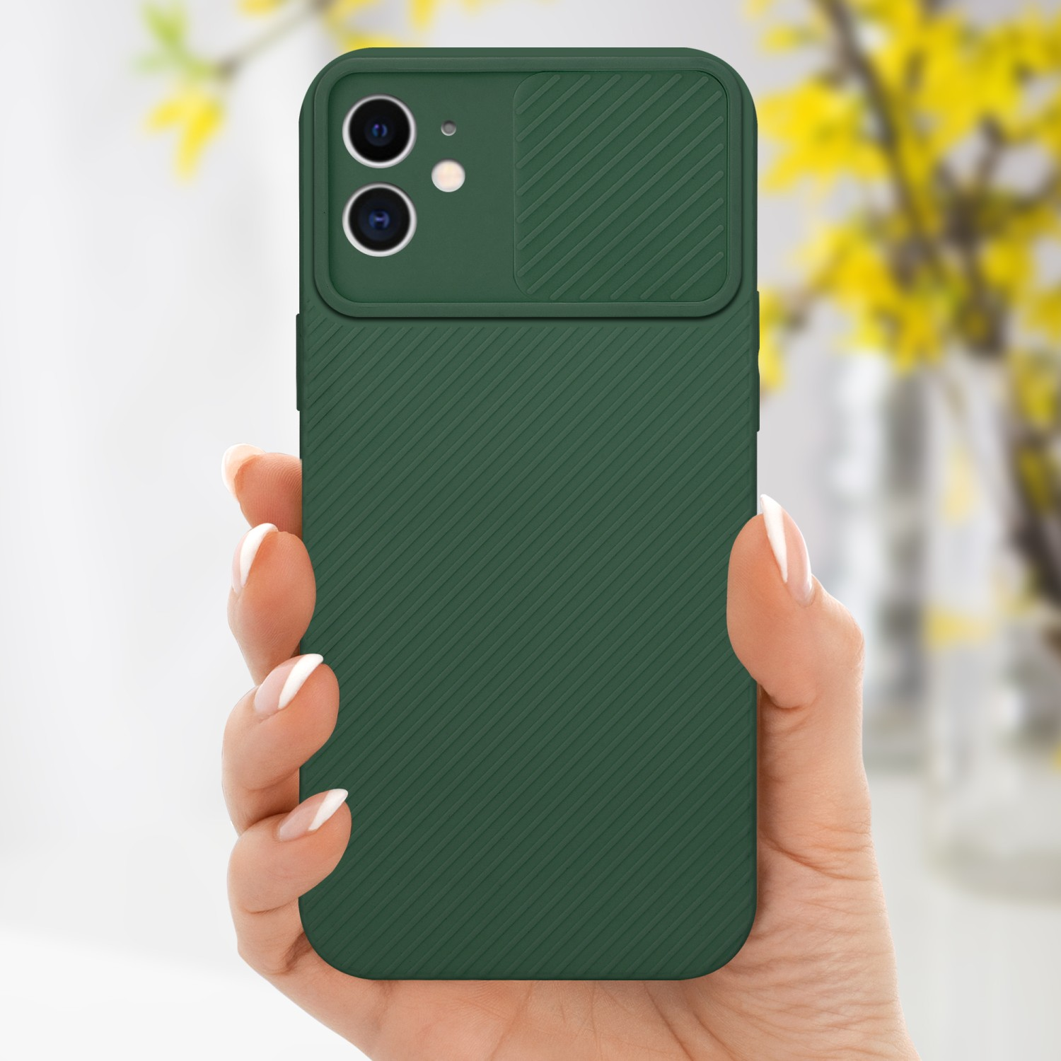 Custodia verde per telefono con copertura per fotocamera, tenuta da una persona. Fiori gialli sullo sfondo.