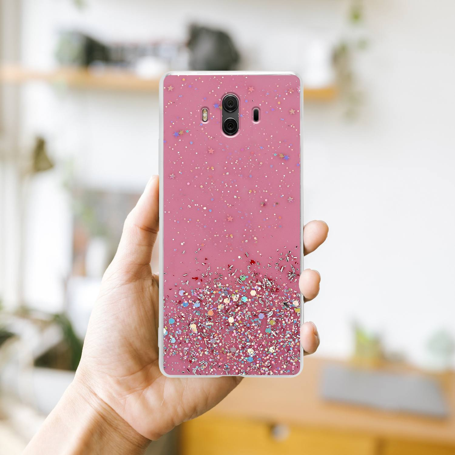 Hand, die eine rosa Handyhülle mit Glitzer hält. Verschwommener Hintergrund eines Raumes.