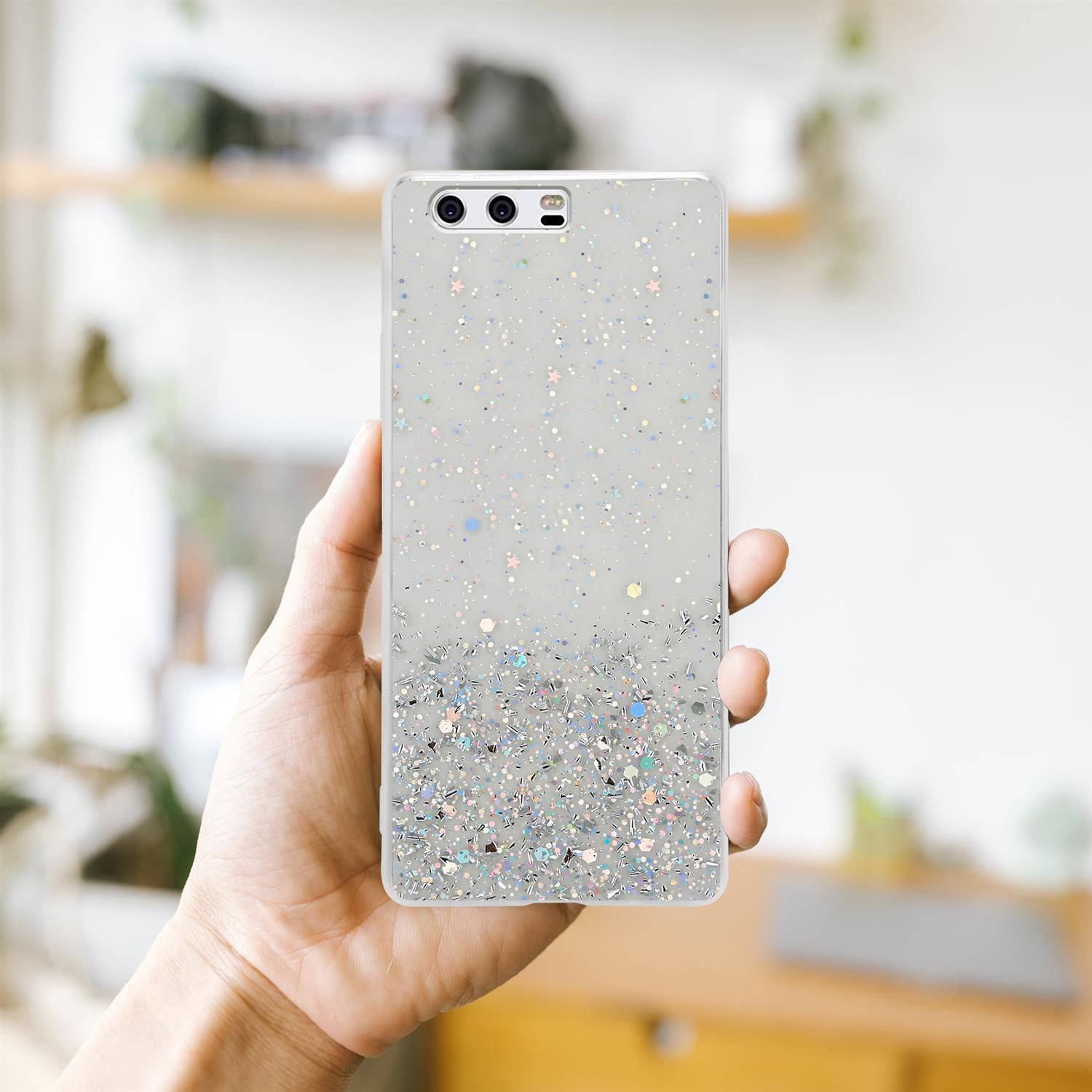 Hand hält eine transparente Handyhülle mit Glitzer. Ein verschwommener Raum ist im Hintergrund.