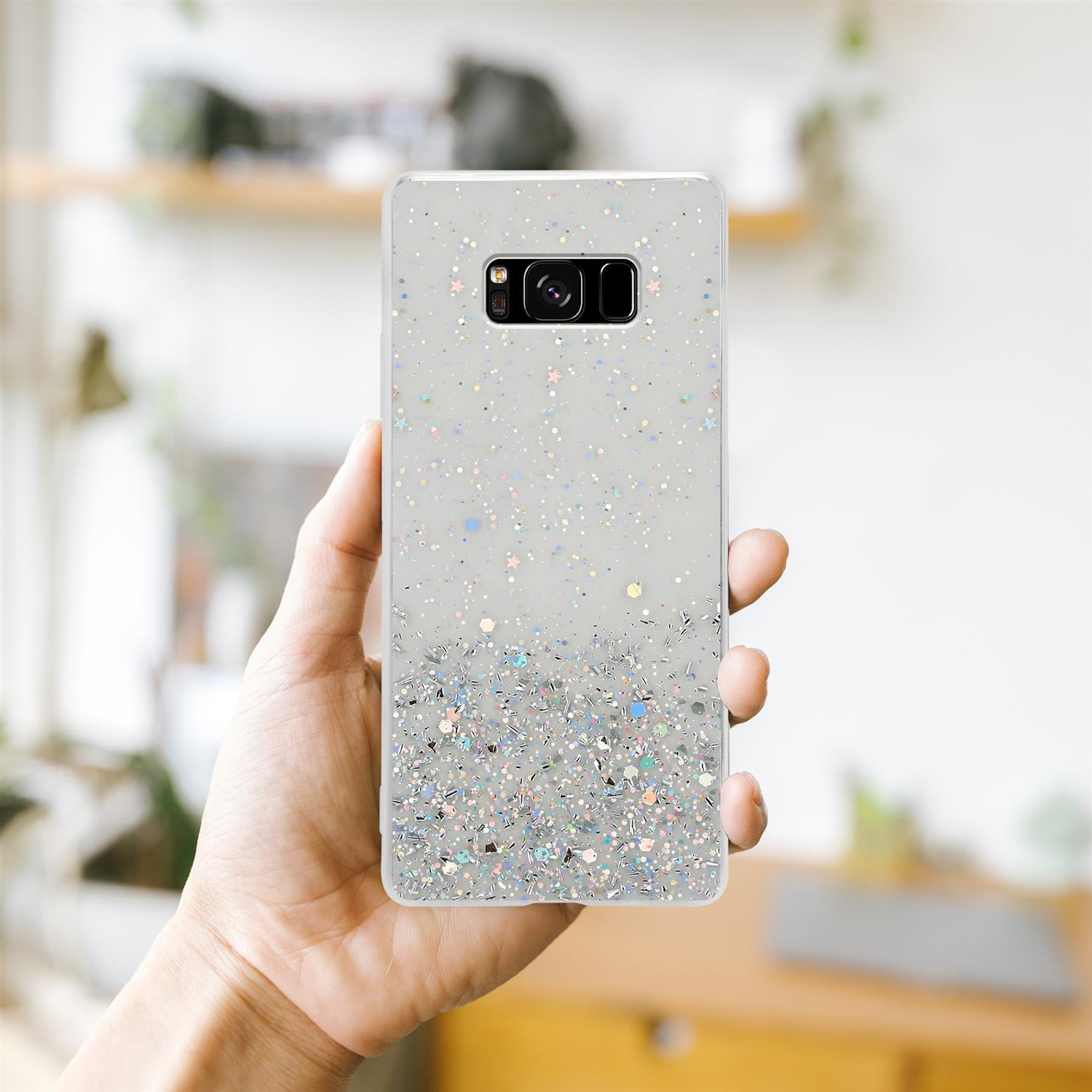 Hand hält ein Telefon mit Glitzer, weiß und silber, vor verschwommenem Hintergrund.
