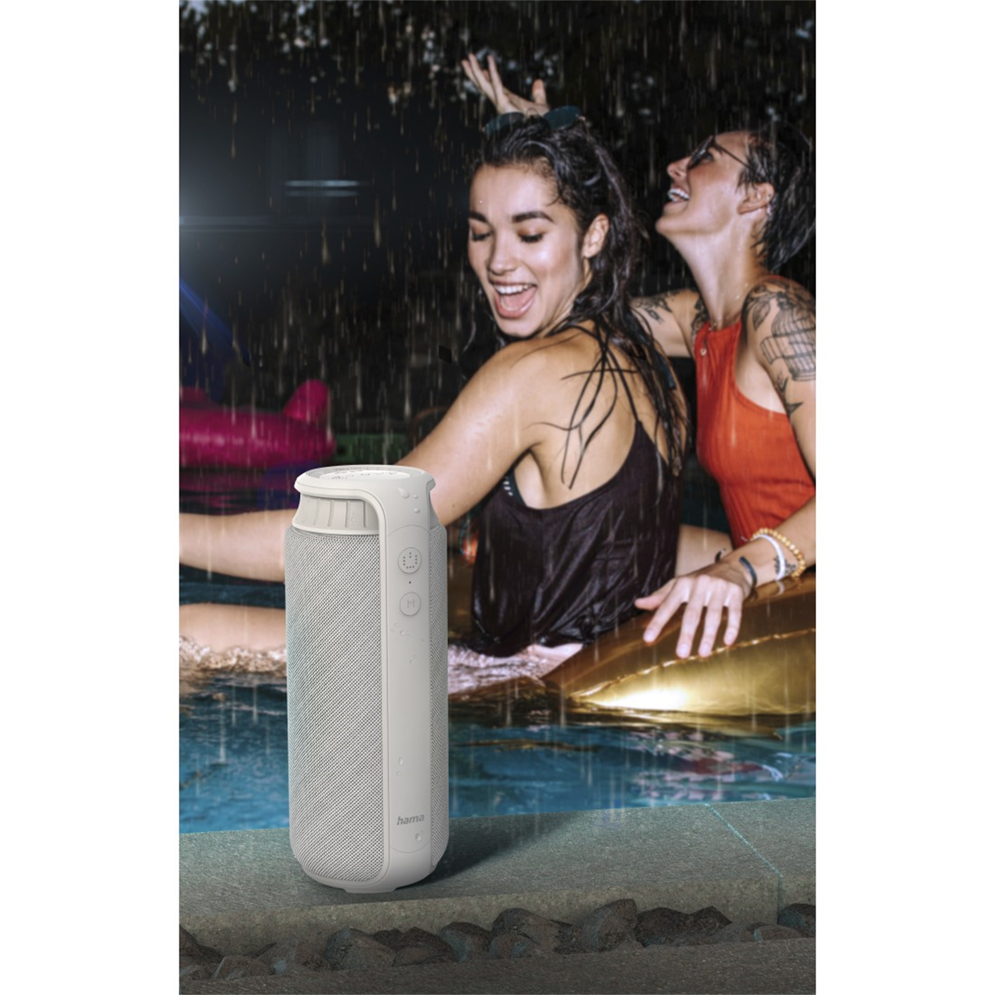 Dwie kobiety w basenie, jedna śmieje się, gdy spada woda. Głośnik stoi na brzegu basenu.