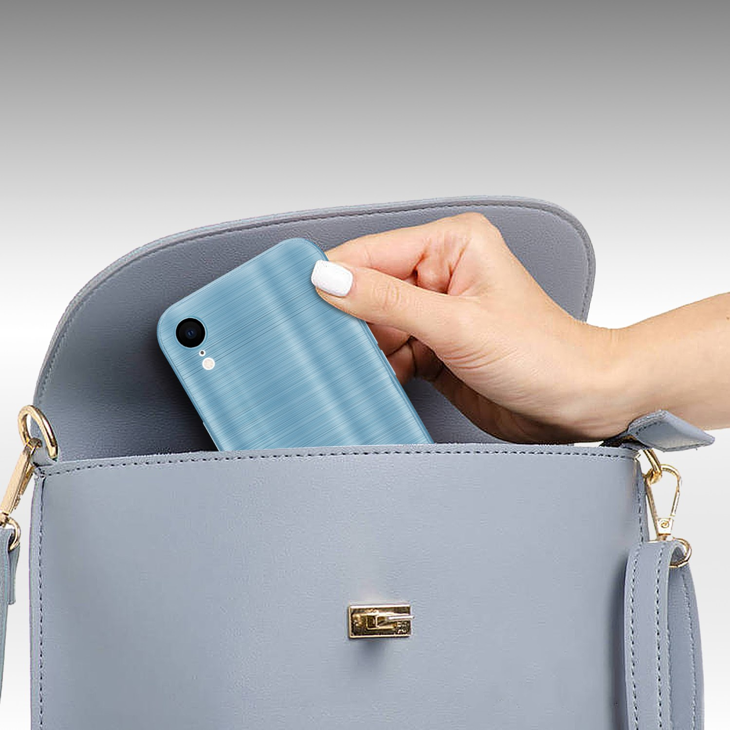 Eine Hand legt ein blaues Telefon in eine hellgraue Handtasche auf hellgrauem Hintergrund.