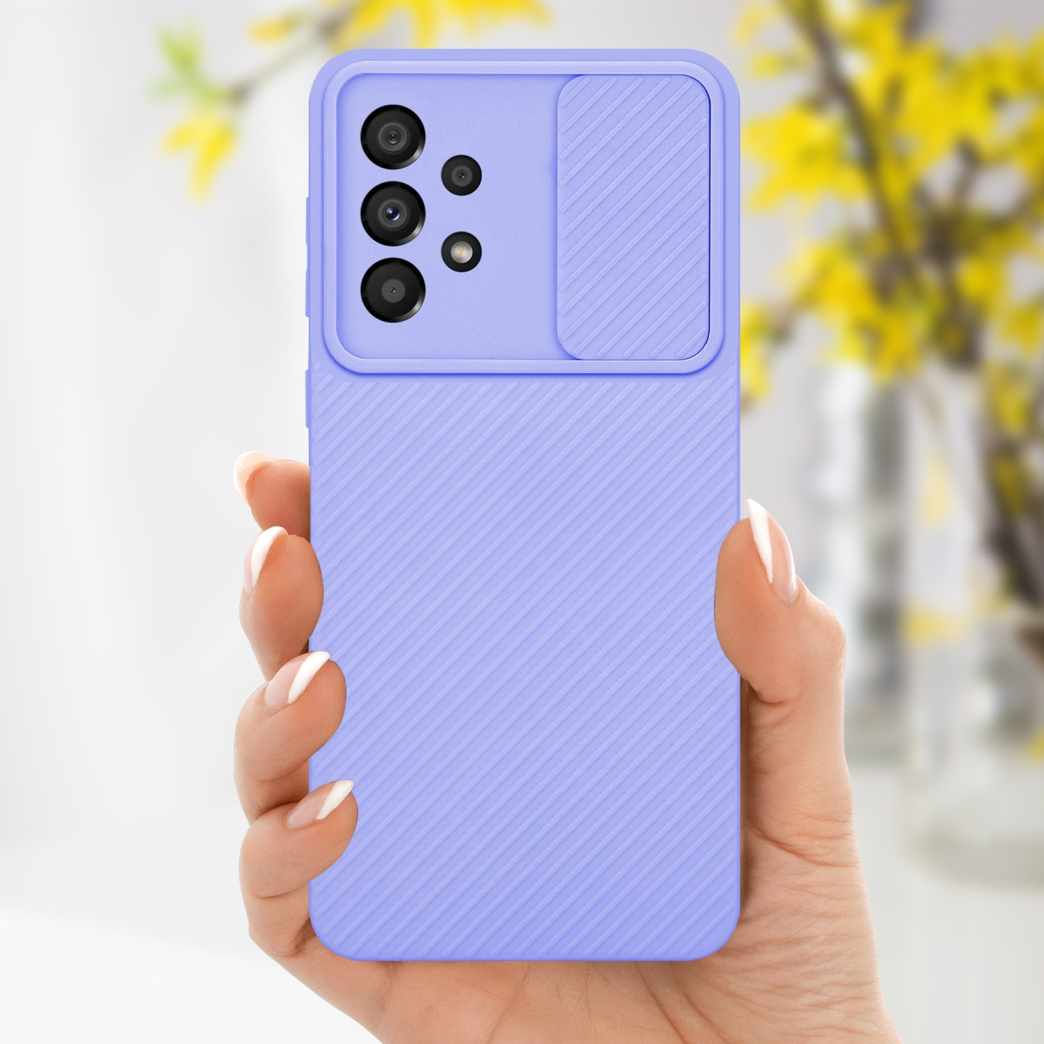 Mano che tiene una custodia lilla con copertura della fotocamera e fiori gialli sfocati sullo sfondo.