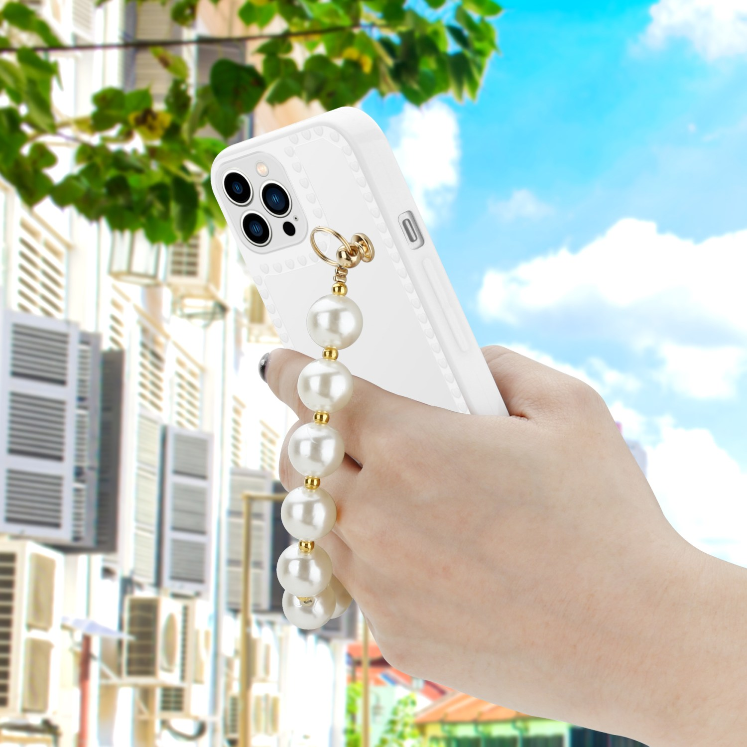 Hand hält weiße Handyhülle mit Perlen-Handgelenkband, Gebäude und Himmel im Hintergrund.