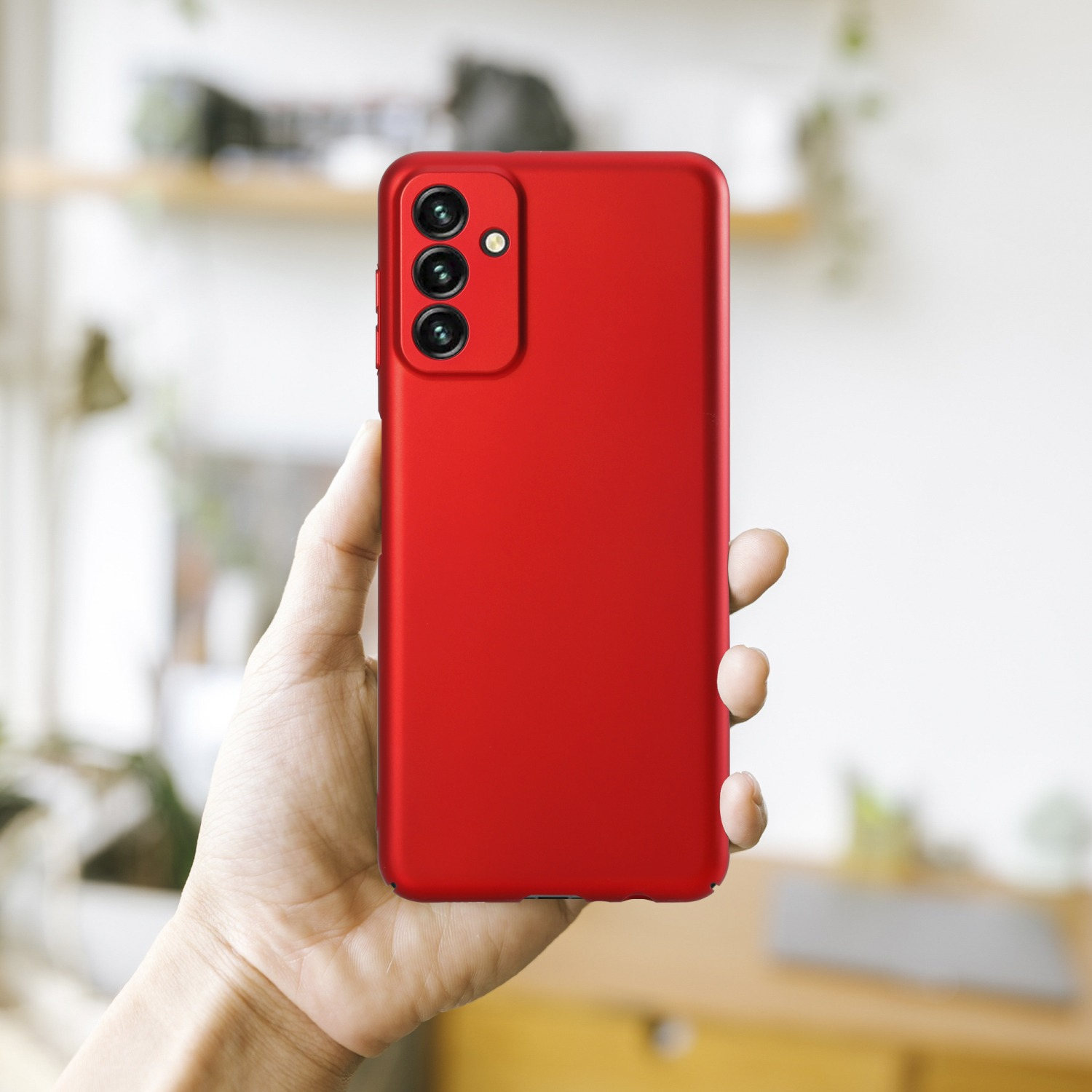 Hand hält ein rotes Telefon mit drei Rückkameras vor verschwommenem Hintergrund.