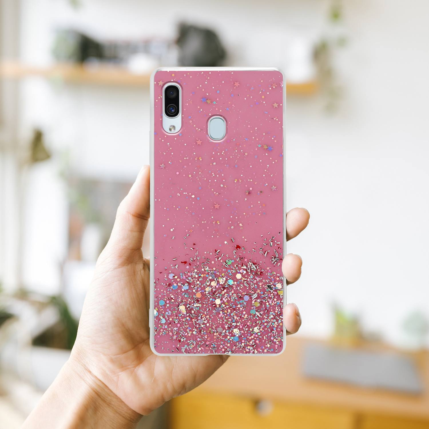 Rosa Glitzer-Handyhülle in der Hand. Der Hintergrund ist verschwommen, was auf eine häusliche Umgebung hindeutet.