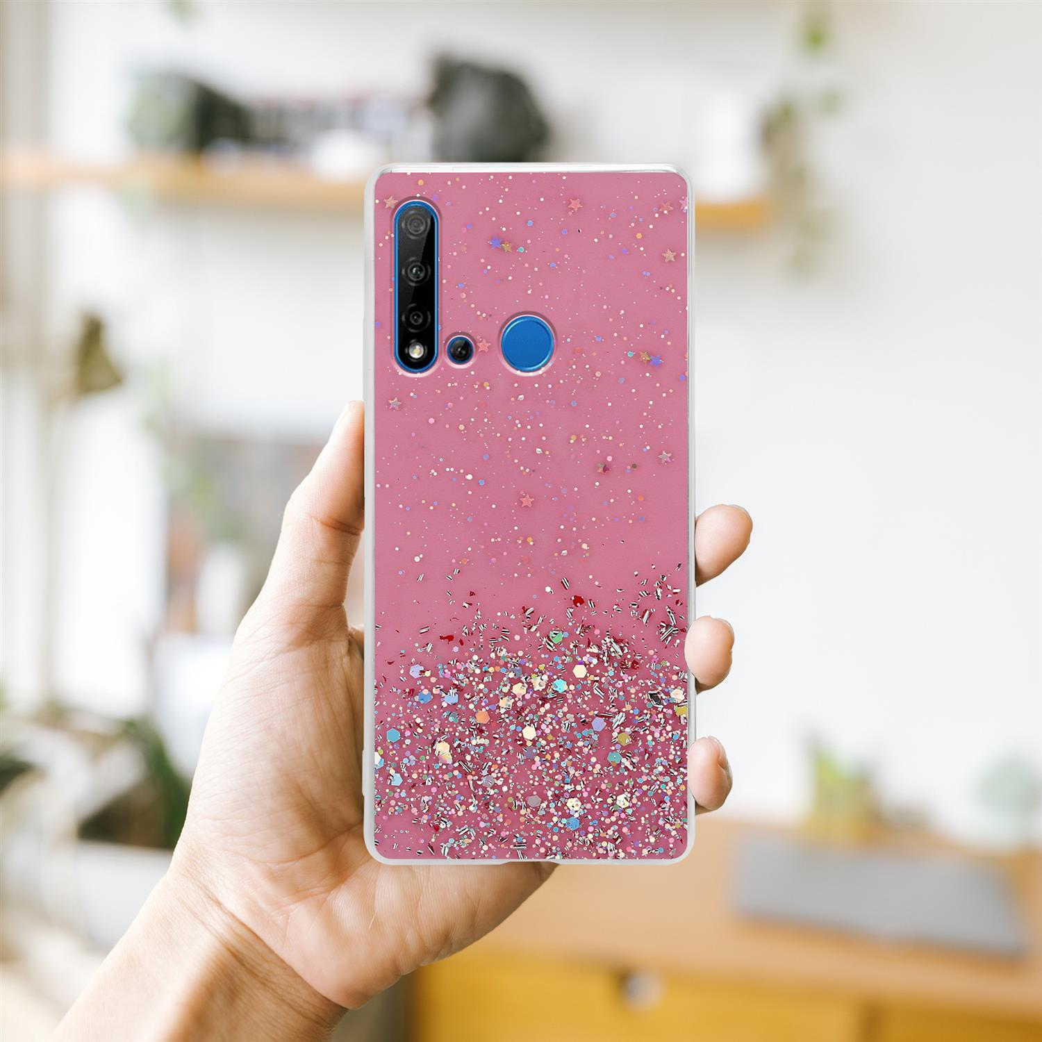 Hand hält pinkfarbene Glitzer-Handyhülle, mit verschwommenem Hintergrund eines Raumes.