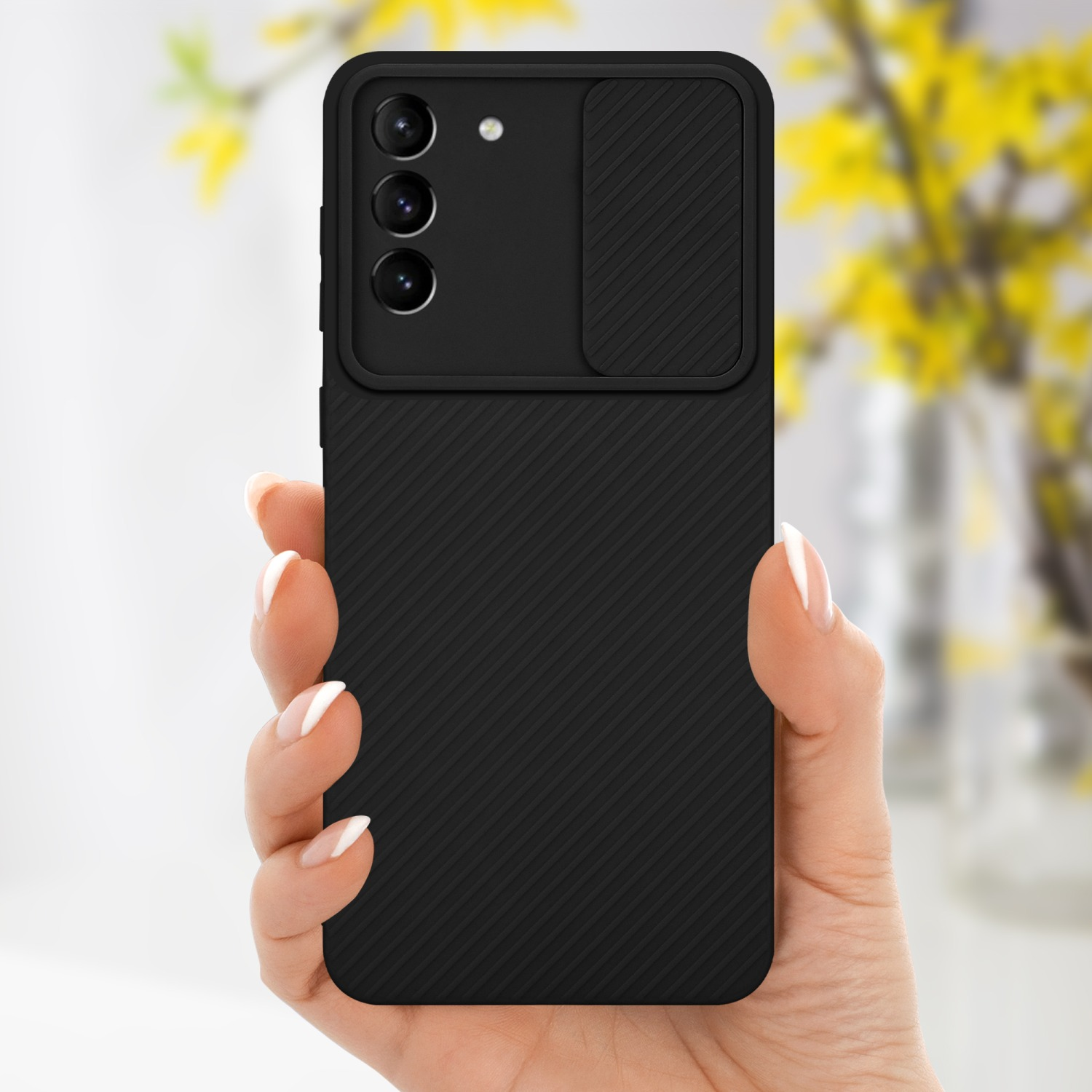 Una mano tiene una custodia nera per telefono con una copertura per la fotocamera. Lo sfondo è sfocato con fiori.