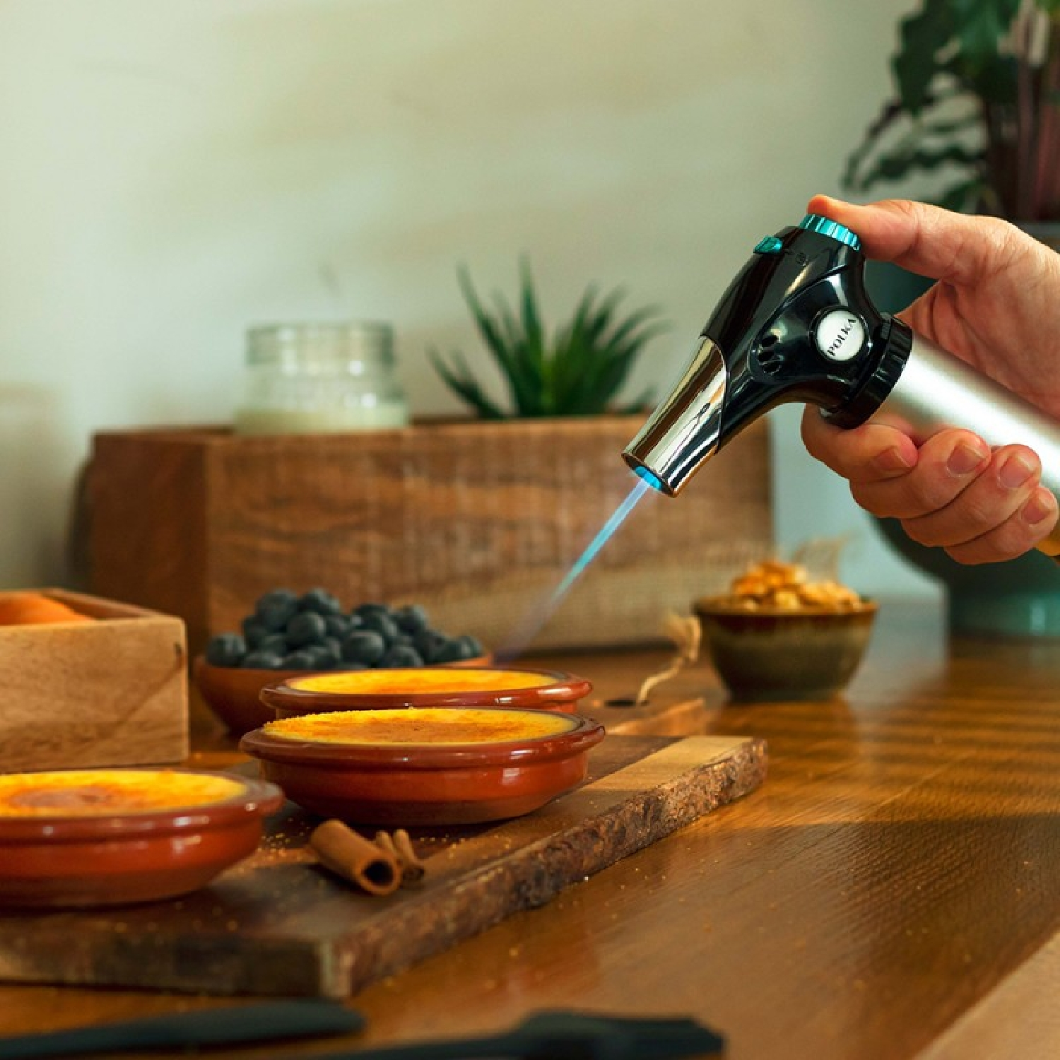Una mano flambea crema catalana. Otros platos, arándanos y una planta en el fondo.