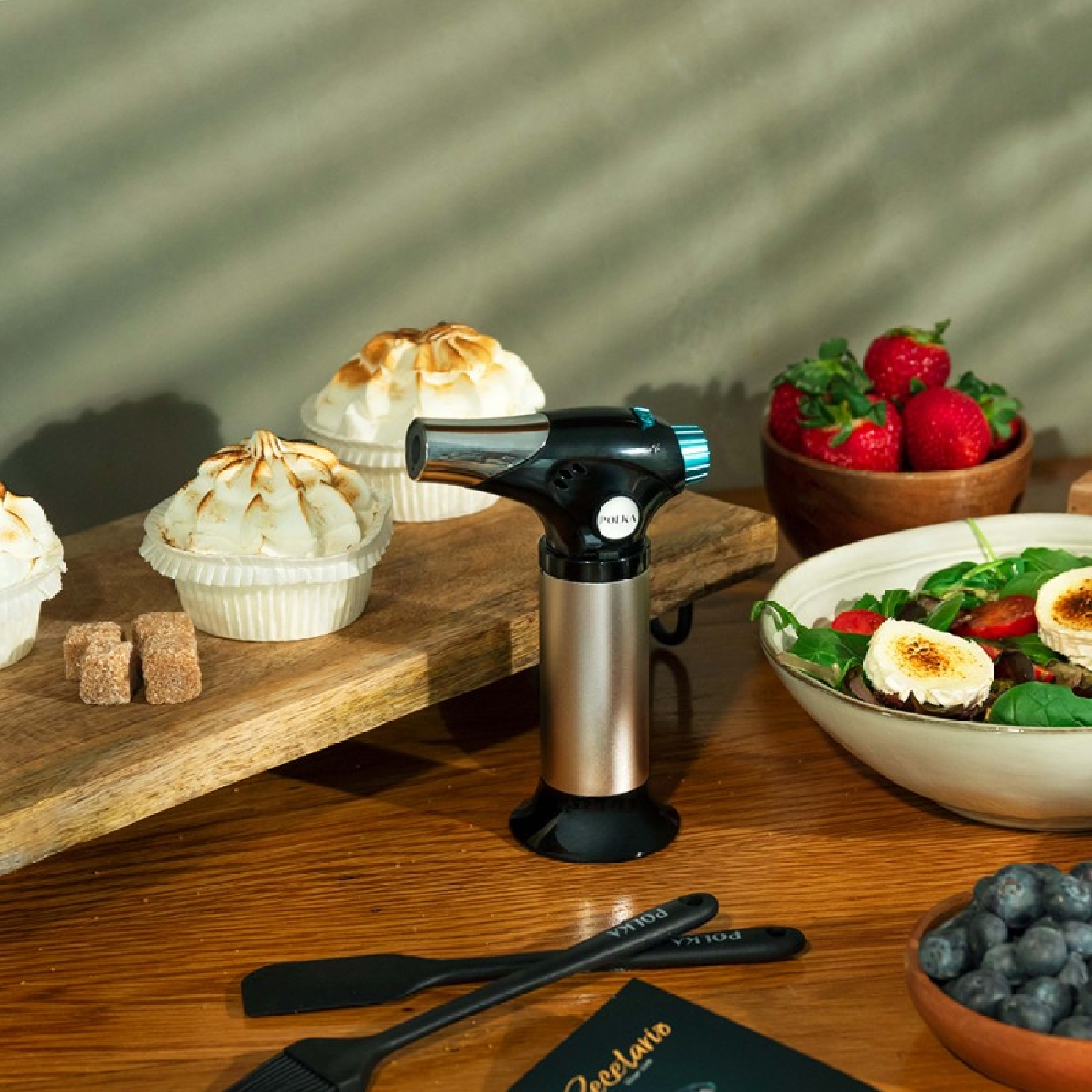 Un soplete de butano junto a cupcakes, fresas y un cuenco de ensalada sobre una mesa de madera.