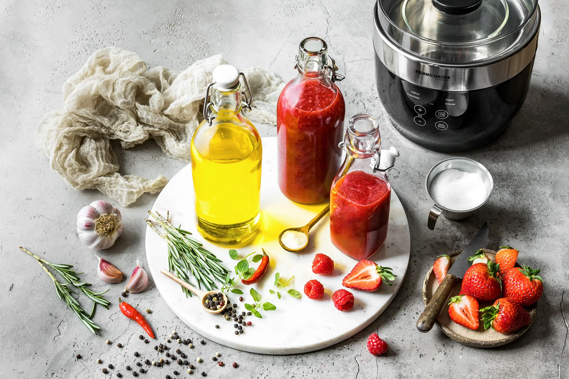 Flaschen mit Öl, Sauce und Inhalt. Kräuter, Gewürze und Erdbeeren auf Marmor. Kochutensilien in der Nähe.