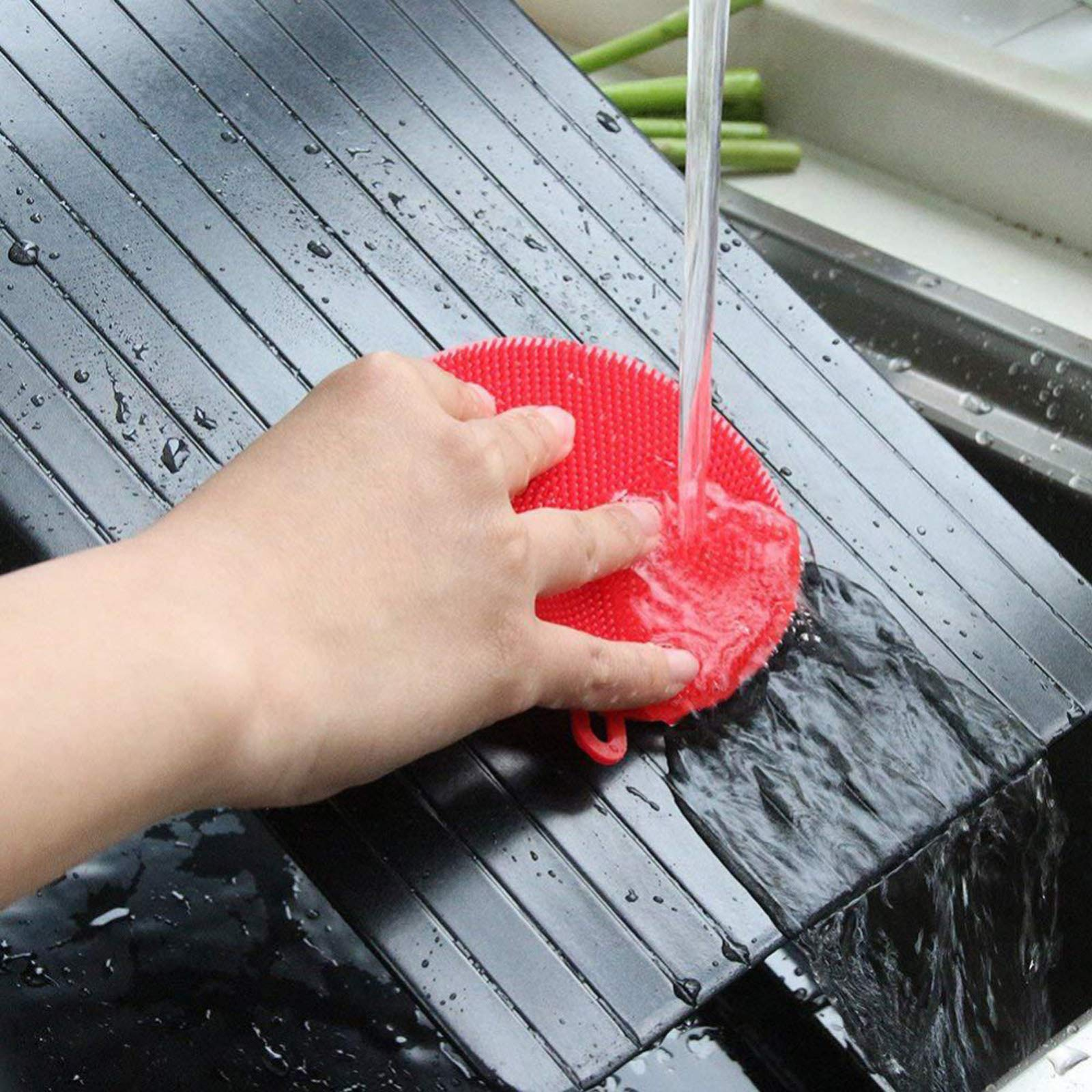 Hand wäscht einen roten Silikon-Geschirrschrubber unter fließendem Wasser in einem Waschbecken.