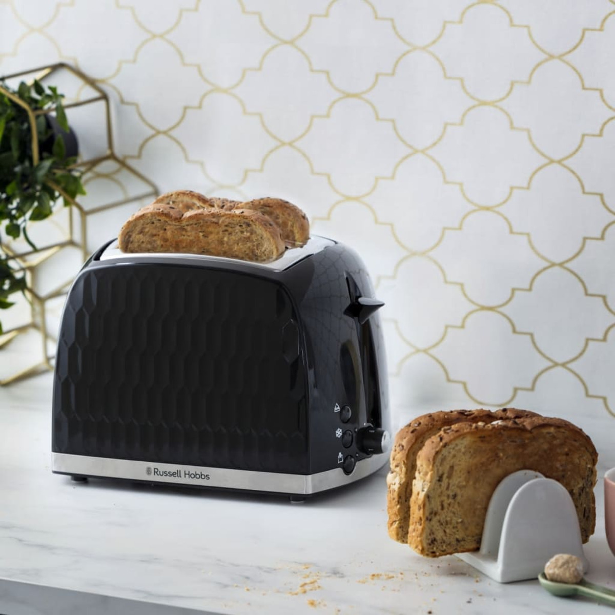 Schwarzer Toaster mit zwei Brotscheiben darin. Zwei weitere Brotscheiben stehen auf einem weißen Ständer.
