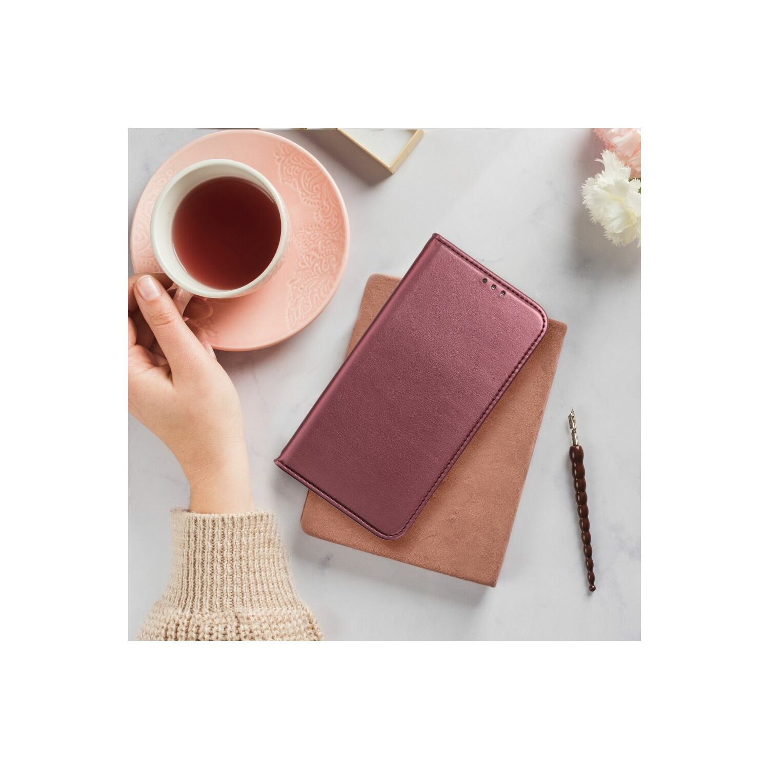Eine Hand hält eine Teetasse auf einer Untertasse, neben einem Notizbuch und einer Handyhülle auf einer Marmoroberfläche.