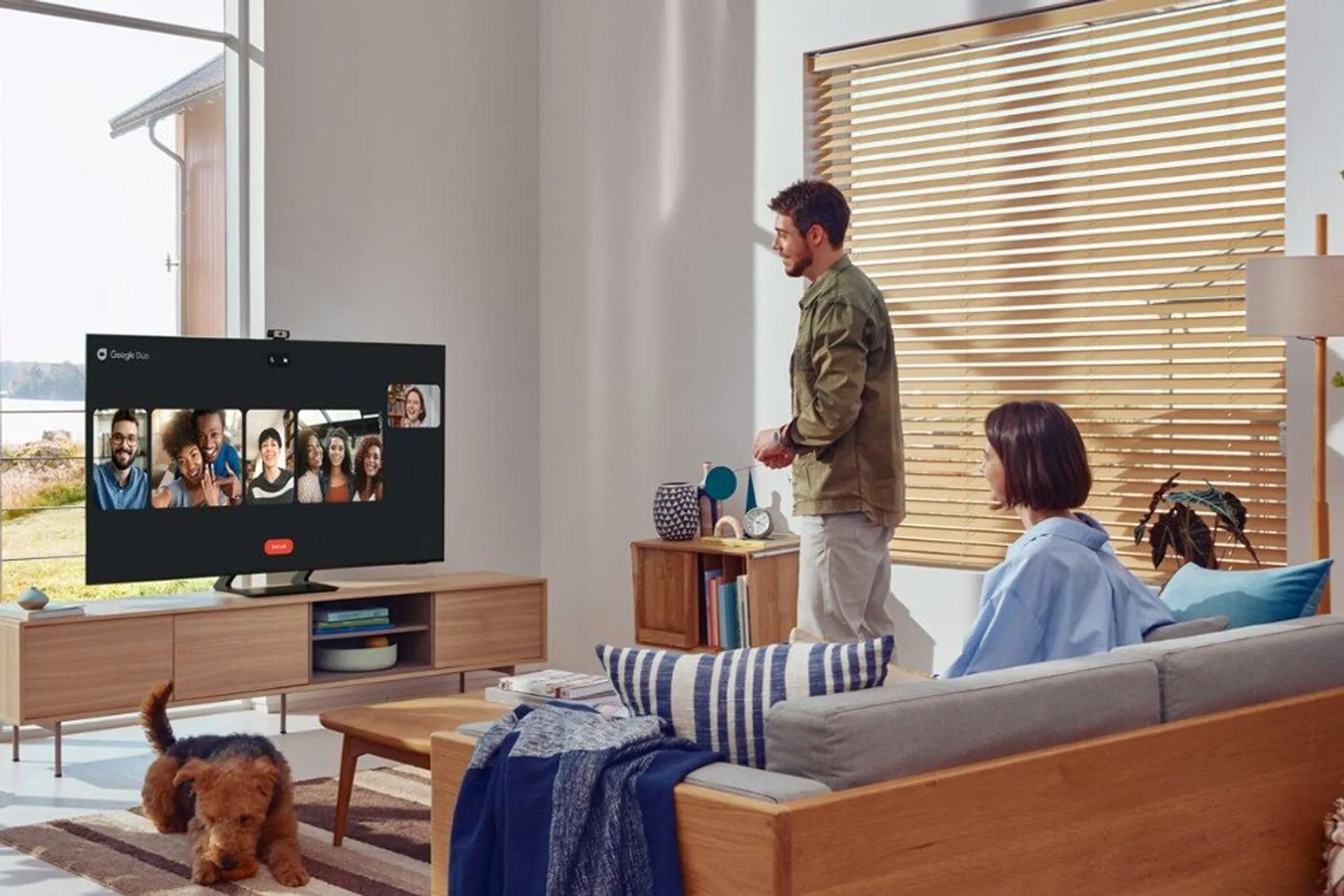 Dwie osoby prowadzą rozmowę wideo na dużym telewizorze. Pies siedzi na podłodze. Scena we wnętrzu.