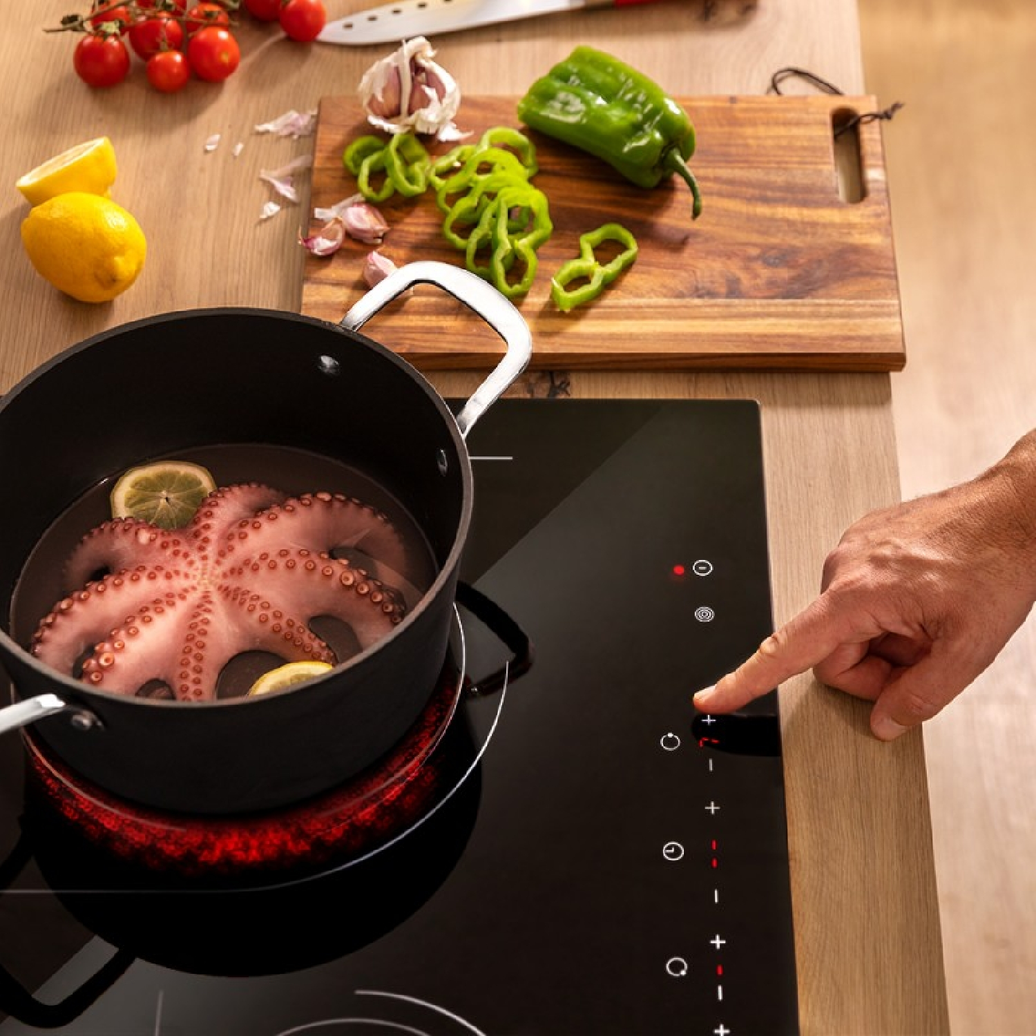 Ośmiornica w garnku na czarnej kuchence. Warzywa i dłoń w kadrze.