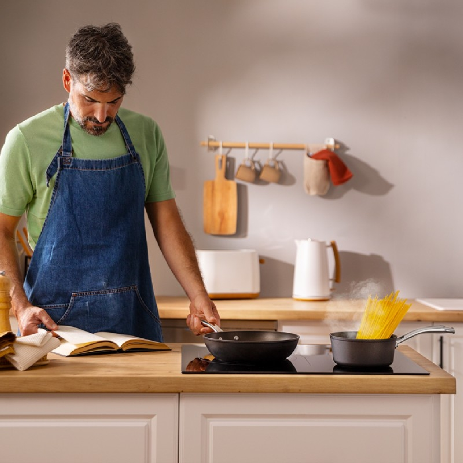 Mężczyzna w kuchni czyta książkę podczas gotowania makaronu na patelni.
