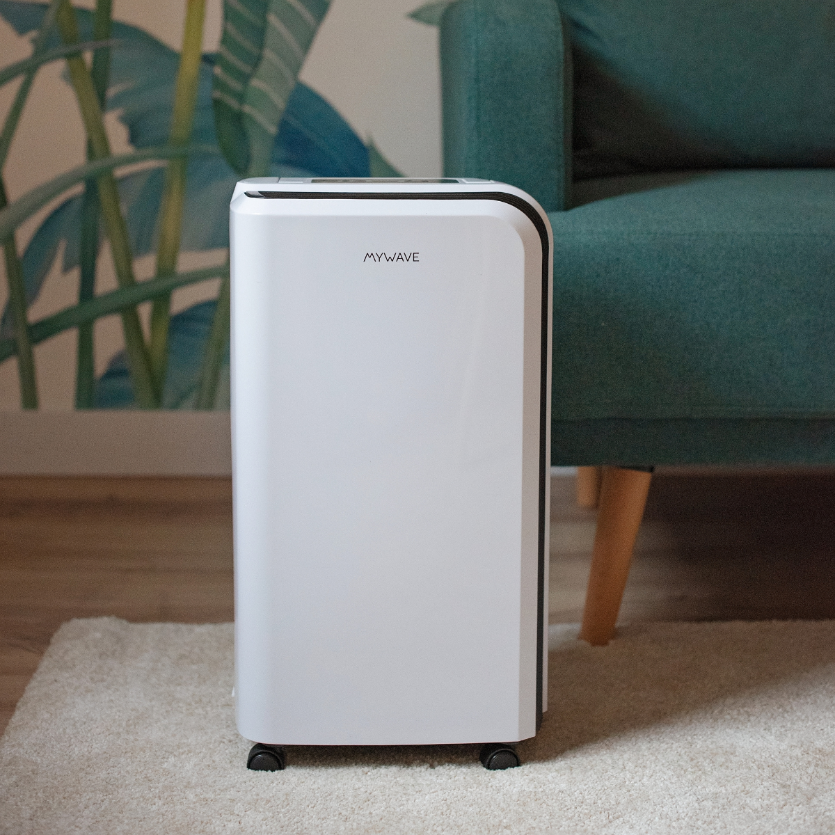 Purificador de aire blanco sobre una alfombra clara. Un sillón turquesa en el fondo. Un cuadro de plantas en la pared.