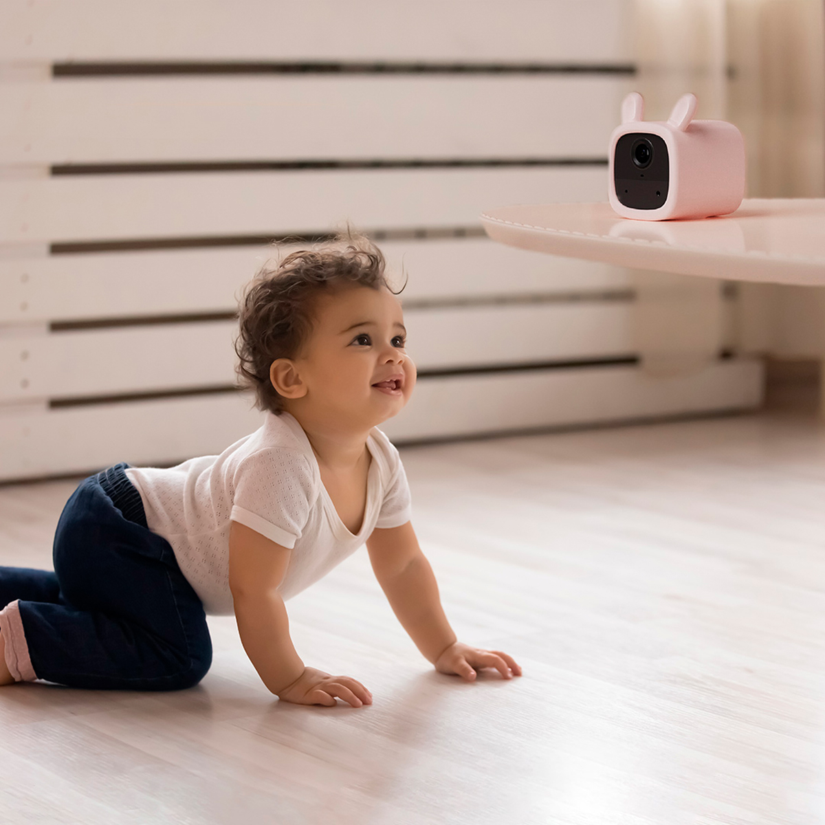 Ein Baby krabbelt auf einem hellen Holzboden auf eine rosa Babyphone-Kamera zu. Das Baby lächelt.