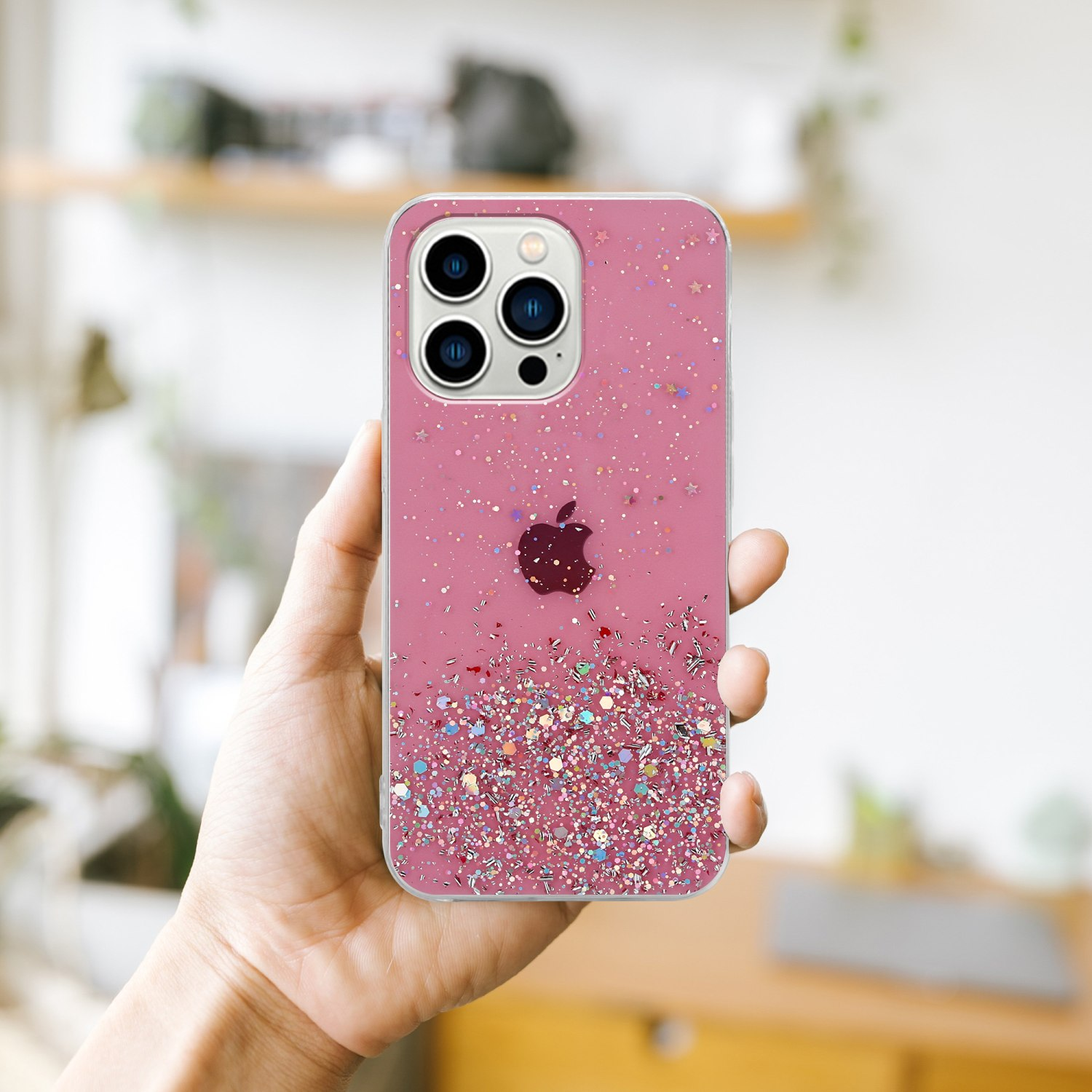 Hand hält ein rosa Telefon mit klarer Hülle und Glitzer. Hintergrund ist eine verschwommene Inneneinstellung.