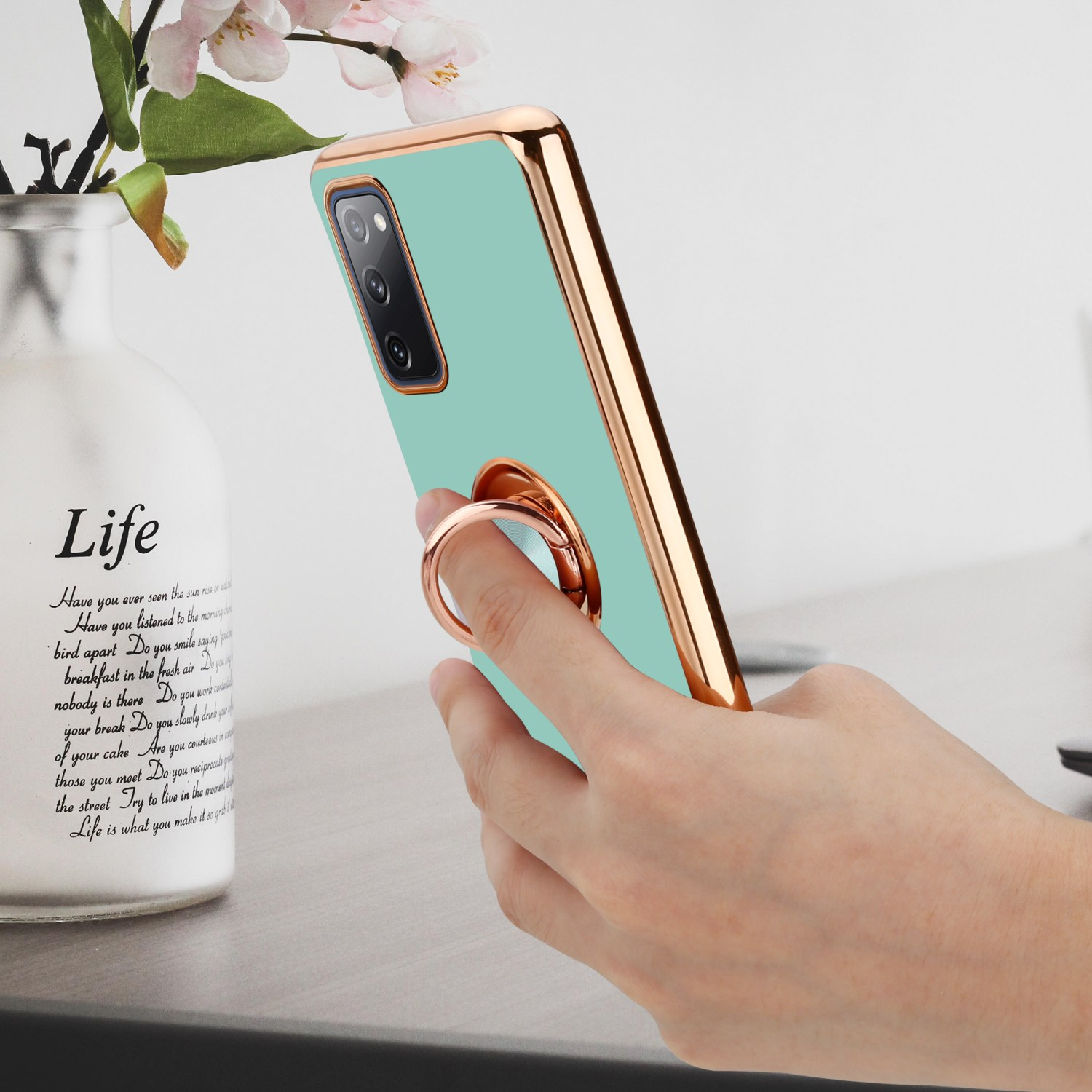 Eine Hand, die ein Telefon mit einer türkisfarbenen Hülle und einem goldenen Rahmen hält. Eine Vase mit Schrift und Blumen ist im Hintergrund.